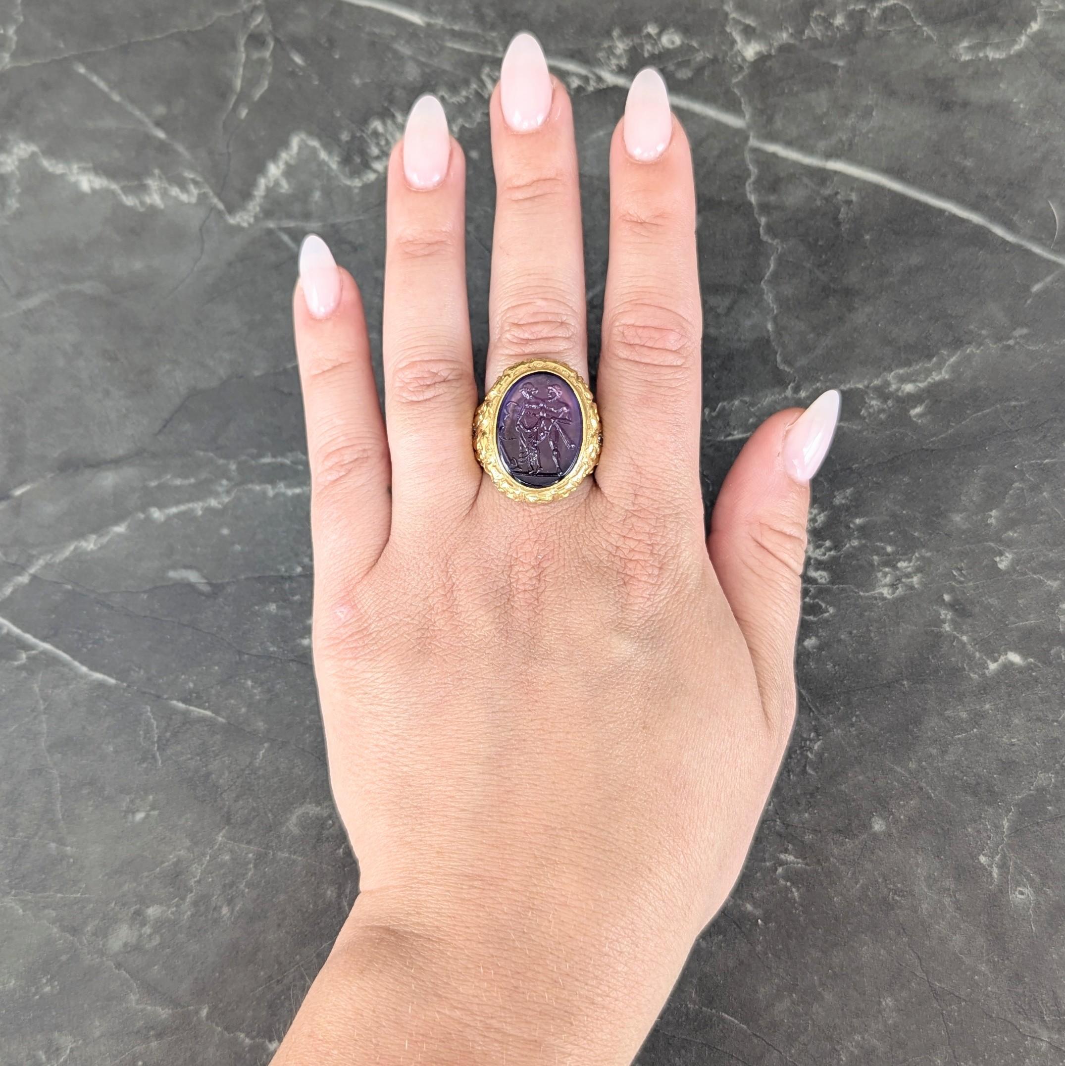 Centering an oval-shaped amethyst measuring 15.0 x 20.0 mm and carved with intaglio
Depicting the Greek myth of Cassandra and Ajax with both figures, an axe, and torch
Bezel set with deeply grooved scrolling floral halo surround and shoulders
Flush
