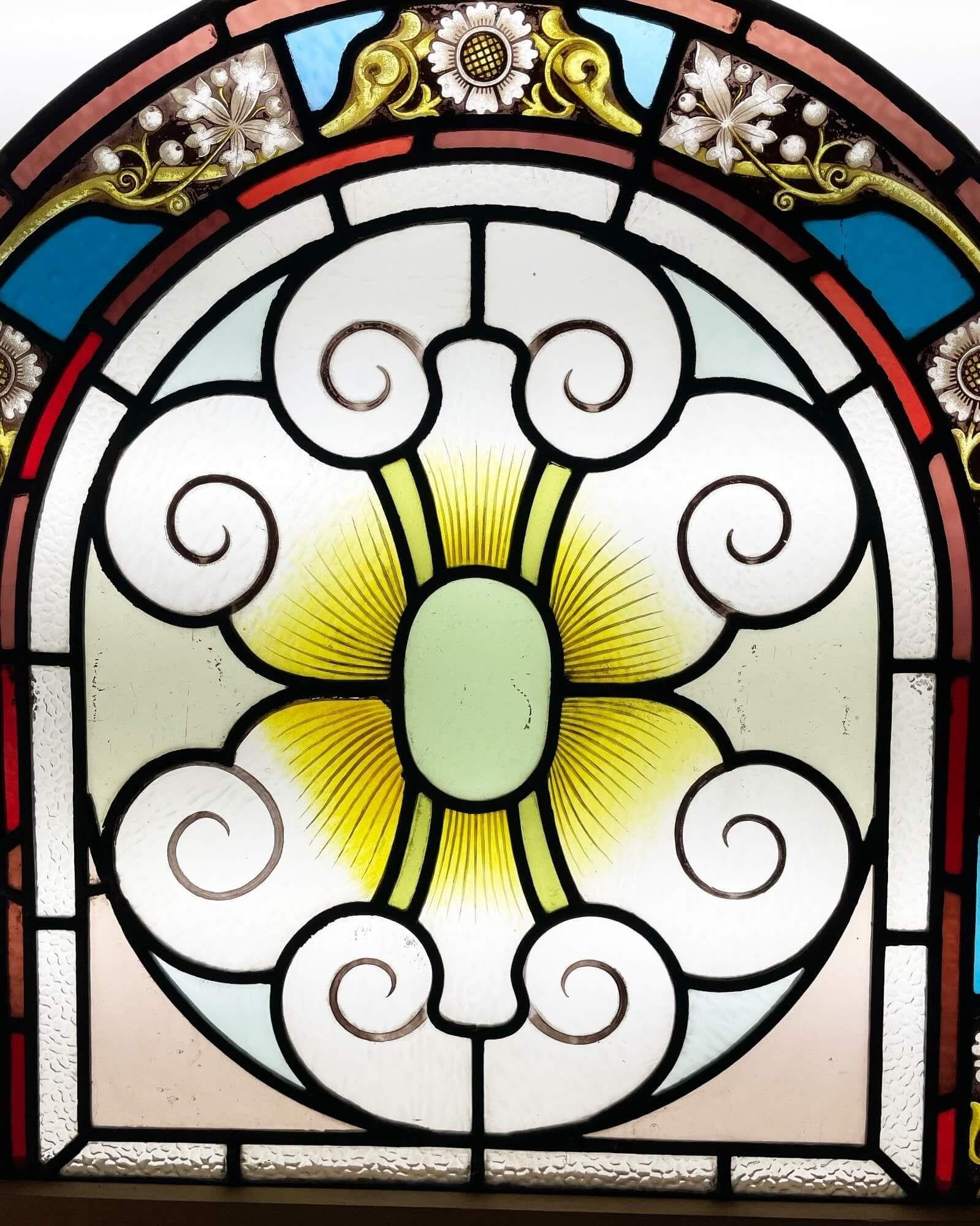 A beautiful antique arched leaded glass window panel dating from the Victorian era circa 1880. This colourful stained glass window features an understated scroll and sunburst design bordered with various stained glass panels in tones of blue, red