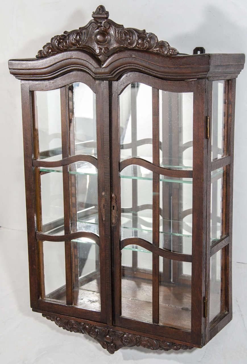 Antique wall mounted curio cabinet and vitrine in dark wood with hand-carved crown moldings and pediments.  Cabinet has mirrored back and two adjustable glass shelves, as well as glass door fronts and glass sides, fitted with exquisite carved wood
