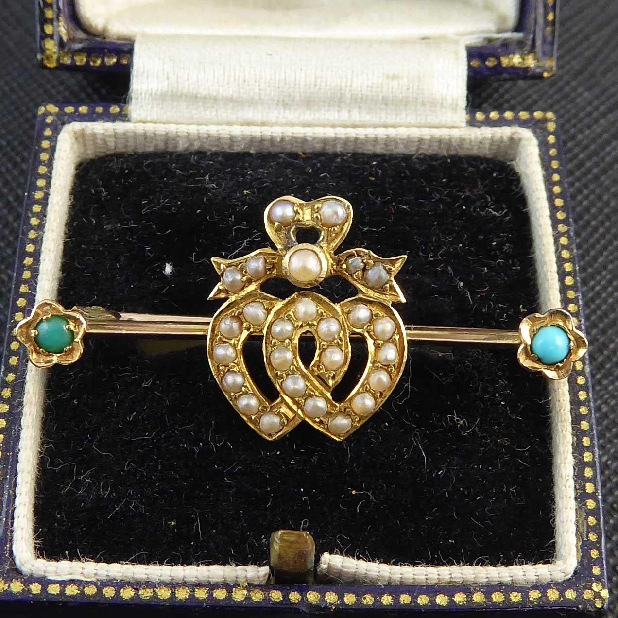 A charming and romantic brooch dating from the Victorian era featuring two pearl studded hearts entwined on a yellow gold knife edge bar.  The top of the hearts a crowned with a pearl ribbon and there are cabochon turquoise set flowers as finials to