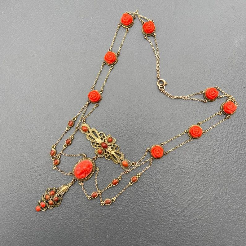 Die bezaubernde antike Halskette stammt aus dem späten 19. bis frühen 20. Jahrhundert und zeigt ein atemberaubendes Design aus Korallen-Zelluloid-Kameen und Korallen-Glascabochons in einem verzierten goldfarbenen Metallrahmen. Die Stücke sind mit