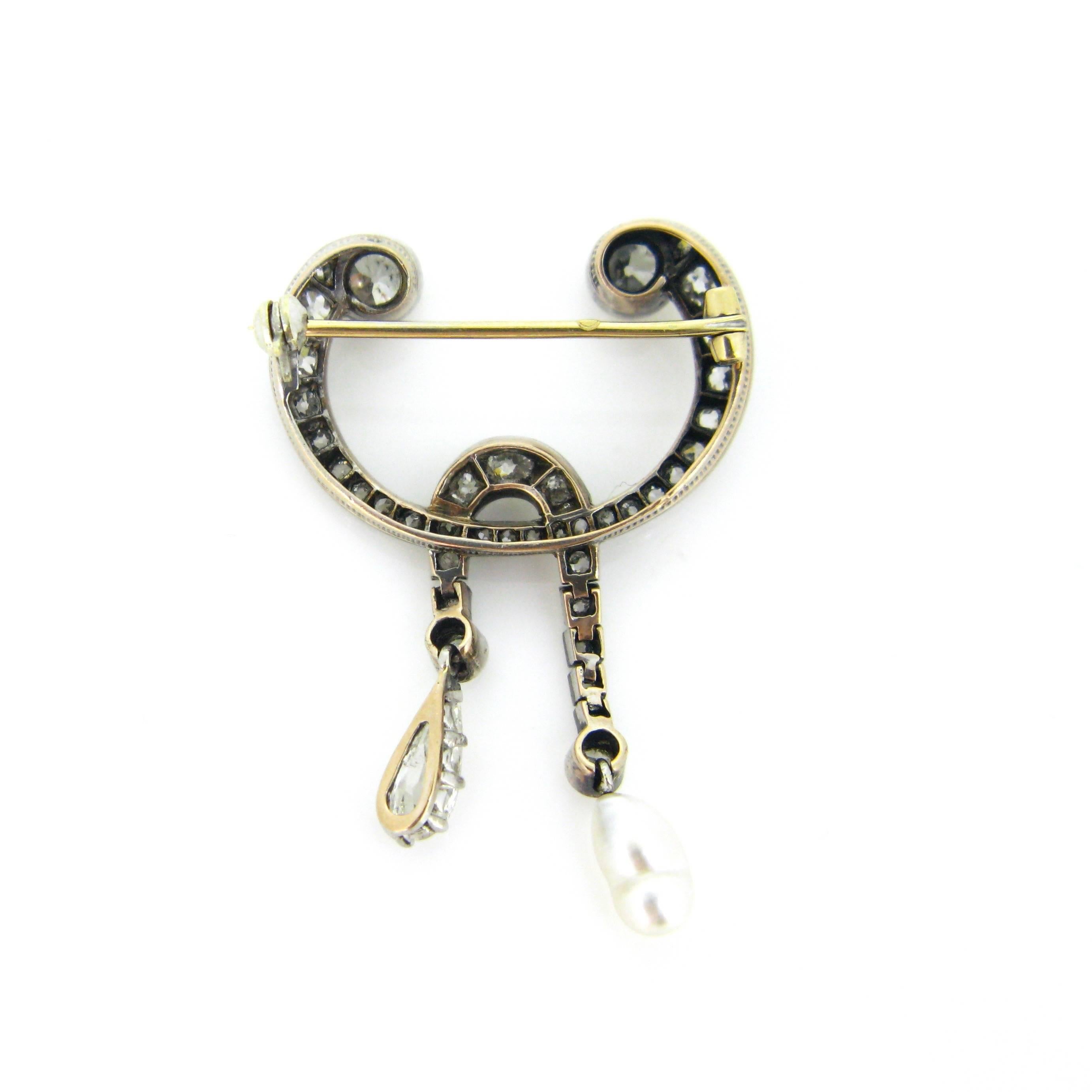 A ravishing and very unique brooch. This one has wonderful curving shapes. It features two sparkling old mine cut diamonds on the top and it ends on 2 dangling parts : on one side we have a natural baroque pearl and on the other side we have a