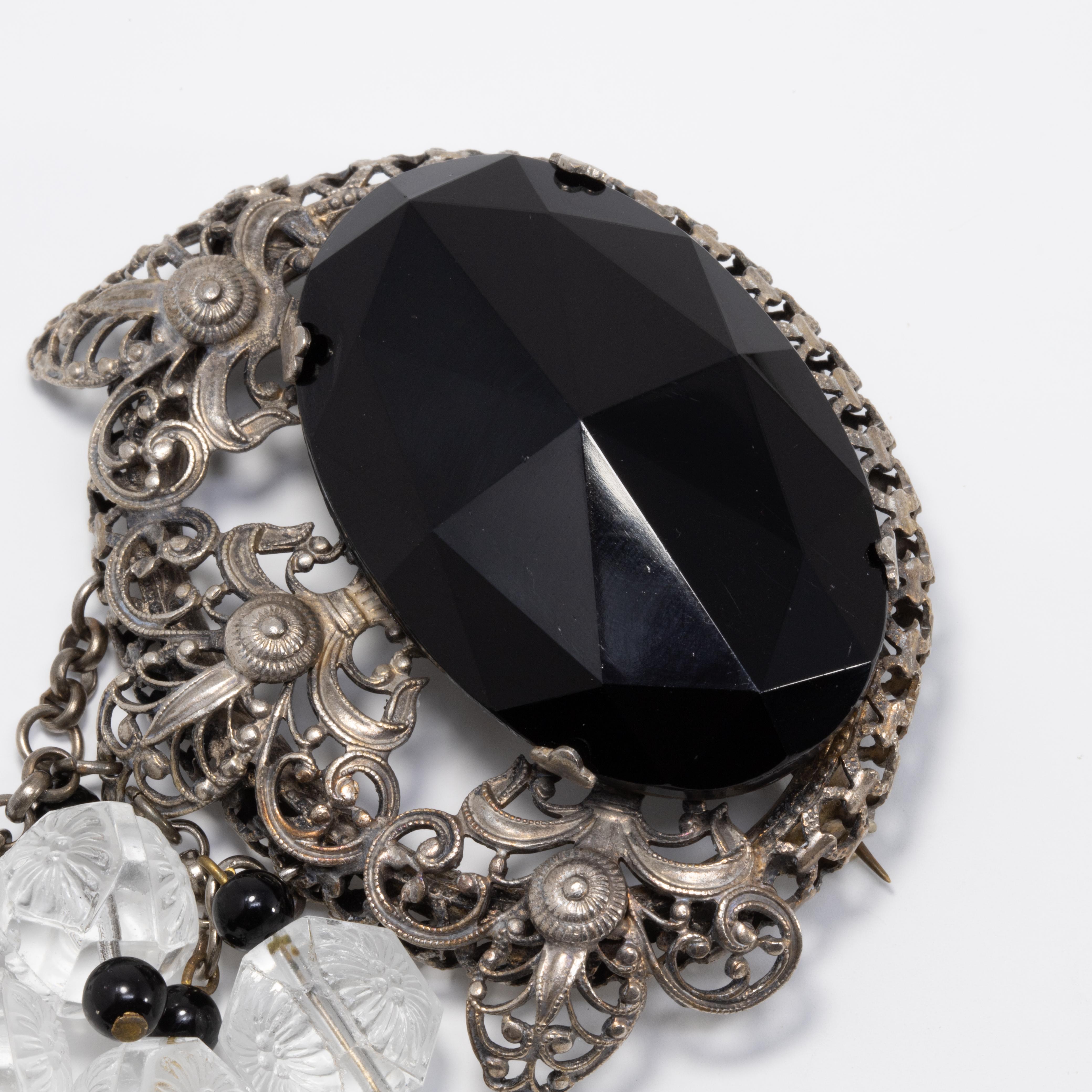 A large, bold, ornate Victorian brooch featuring a large black faceted crystal, prong set in a silvertone setting decorated with Victorian motifs. Four chain strands hang off the bottom, accented with carved clear gemstone crystals and onyx beads.