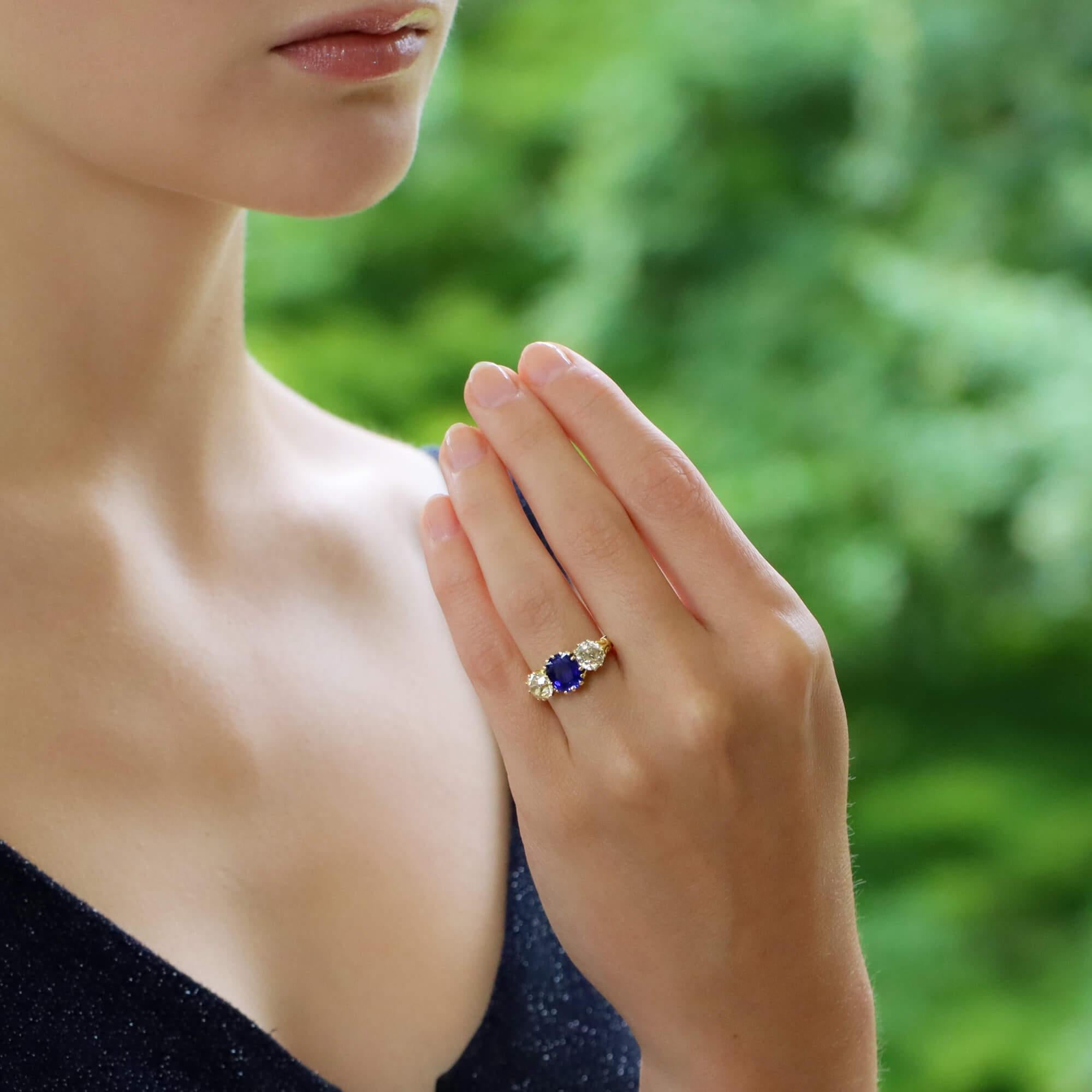 A Victorian sapphire and diamond three-stone engagement ring set in 18 karat yellow gold. 

The ring centrally features an elongated cushion-cut cornflower blue sapphire set with multiple yellow gold claws. The sapphire is then flanked on each side
