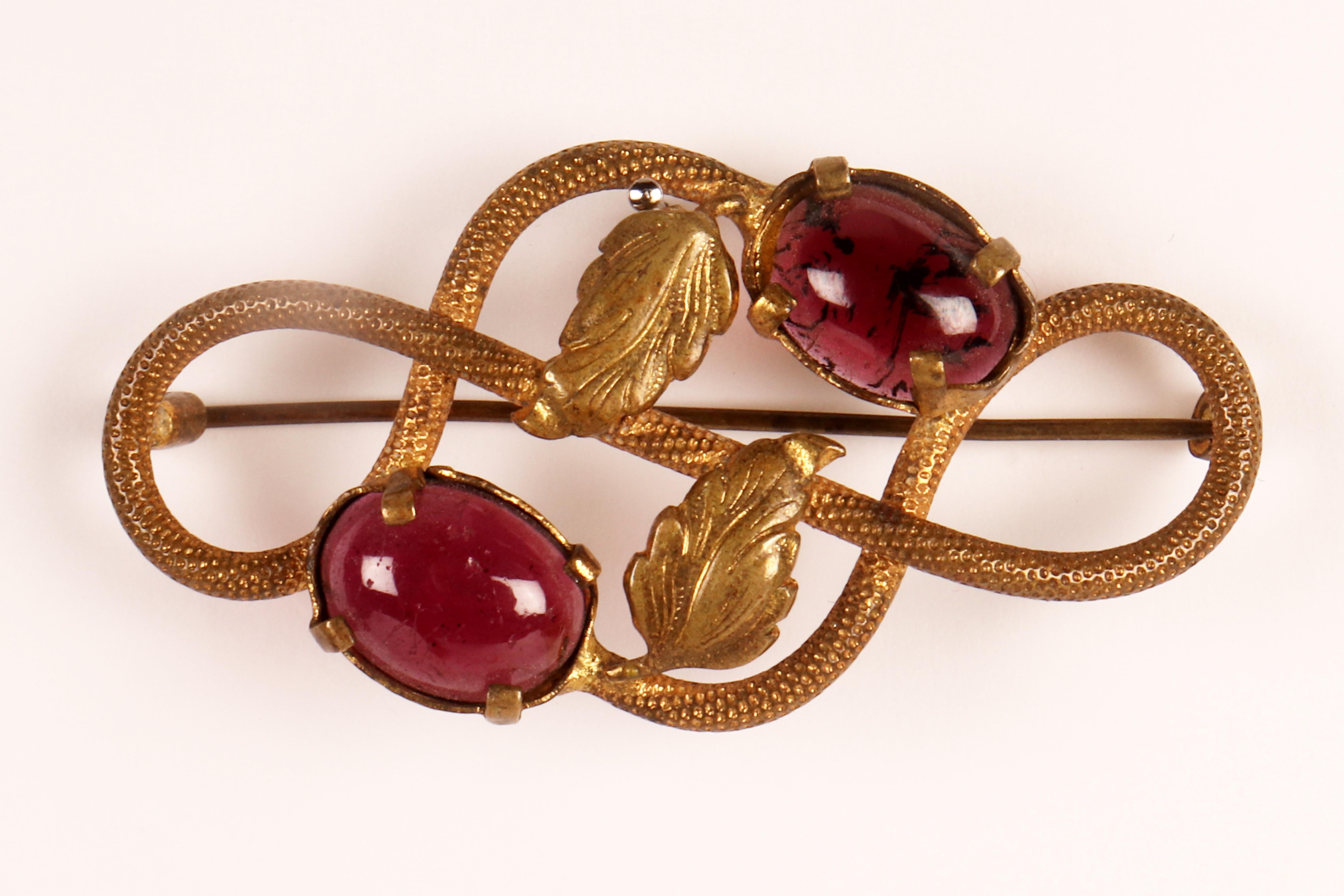 The brooch is made of 14 Kt gold, up of a knot of racemes, two leaves, and two fruits arranged symmetrically in a mirrored knot. The body of the raceme is entirely chiseled with spiral decoration, the leaves finished as a burin. The two oval garnets