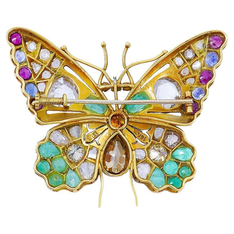 A colorful butterfly pin brooch made of 18k gold, featuring various gemstones, and accented with colored enamel. 
The gems are mount-flush set in the gold “wings” and bezel set in the butterfly’s “body”. The “head” and “wings” feature rose cut