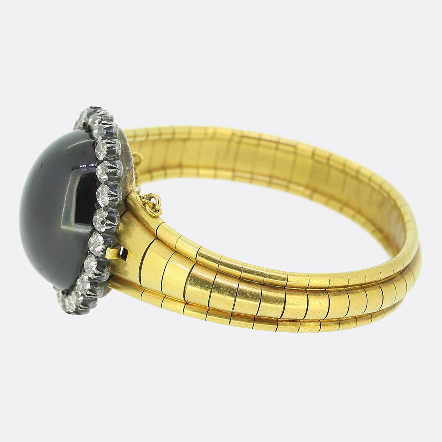 Here we have a beautifully bold bangle come brooch featuring a captivating oval shaped cabochon garnet as the primary stone. This oversized principal stone is surrounded by a halo of chunky old mine cut diamonds, each displaying a brilliant white