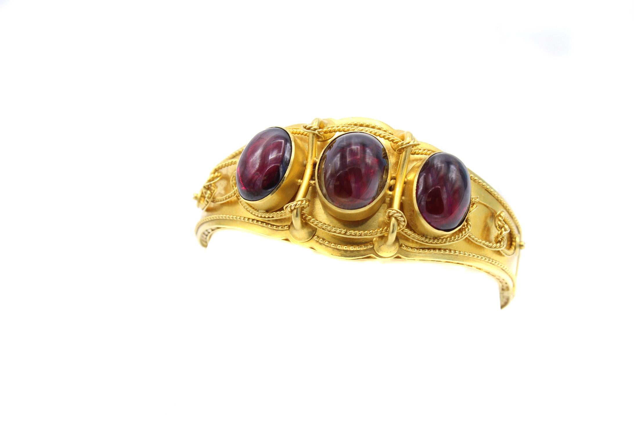 Beautifully designed and masterfully handcrafted, this Victorian gold bangle bracelet features 3 perfectly matched deep burgundy red cabochon garnets. The bangle is worked in a semi-polished gold with detailed braided gold work embellishing the