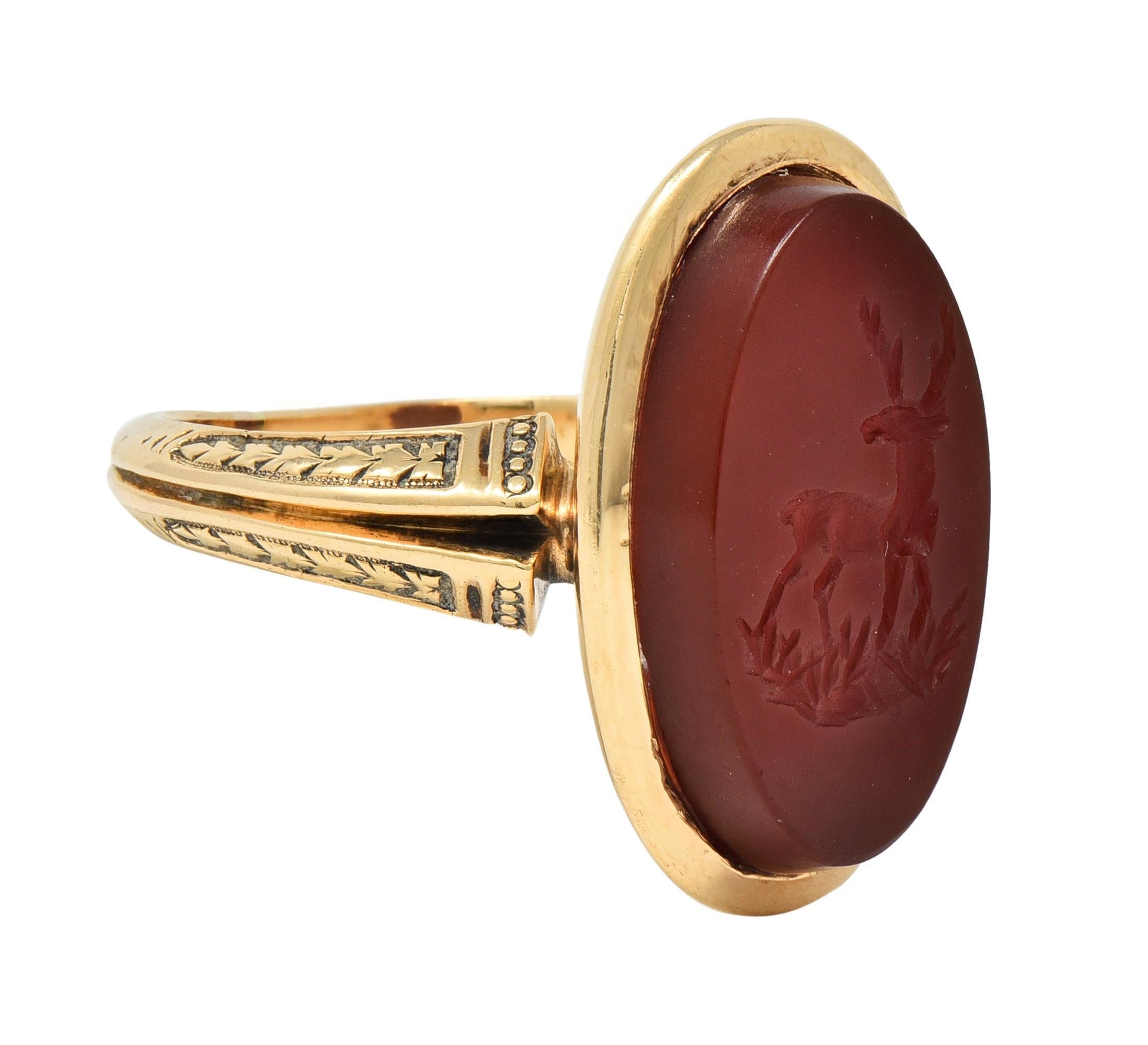 Centering an oval-shaped tablet of carved carnelian measuring 10.0 x 17.5 mm 
Translucent orangey red in color - carved with an intaglio of a stag deer 
With a recessed gold surround flanked by pierced flaring shoulders
Engraved with a decorative