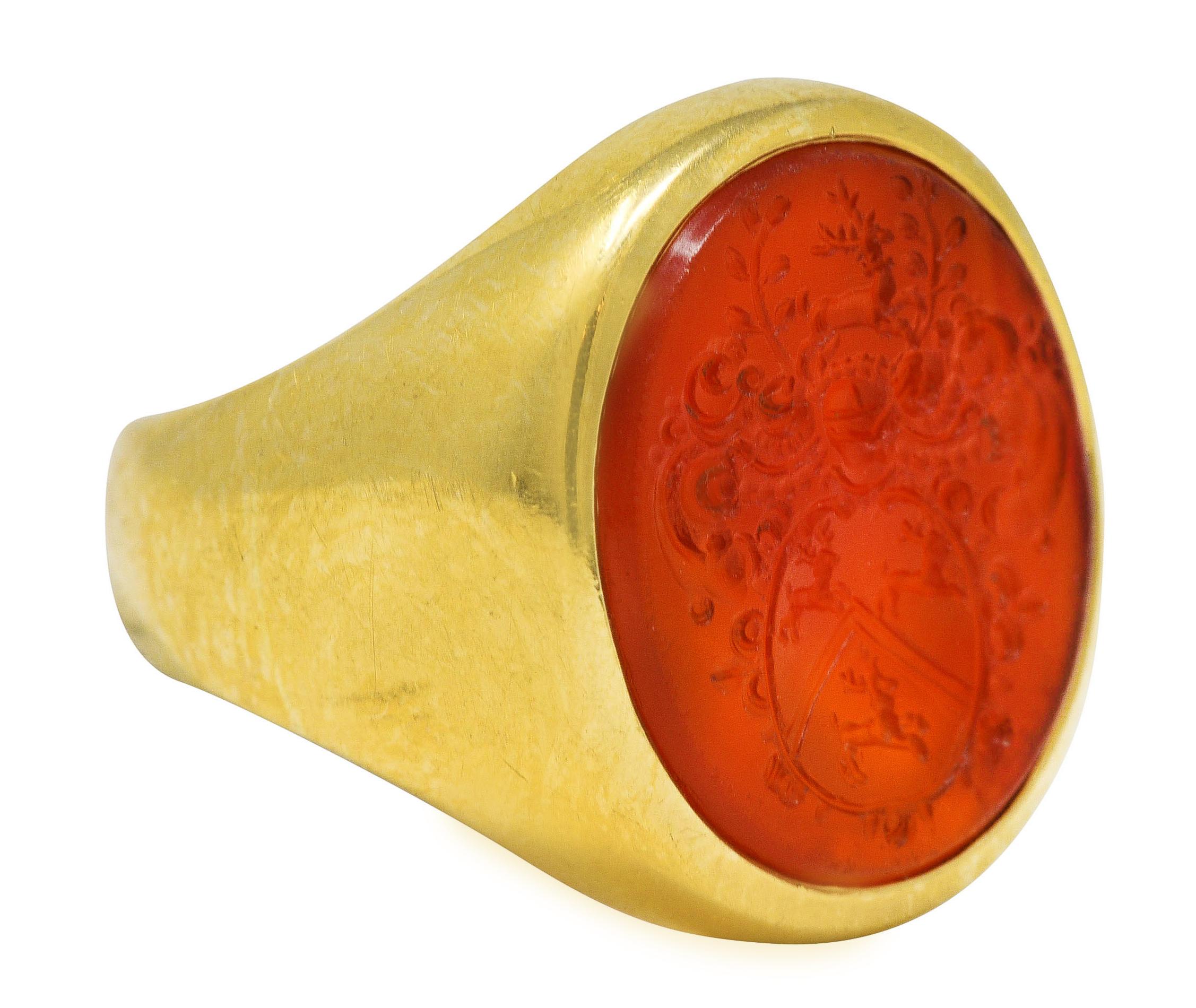 Ring centers a flush set oval carnelian tablet measuring 12.0 x 14.0 mm - translucent medium reddish orange. With intaglio carved to depict the Oxford Jesus College crest - featuring a shield with three stags. Accented by scrolling foliate motifs.