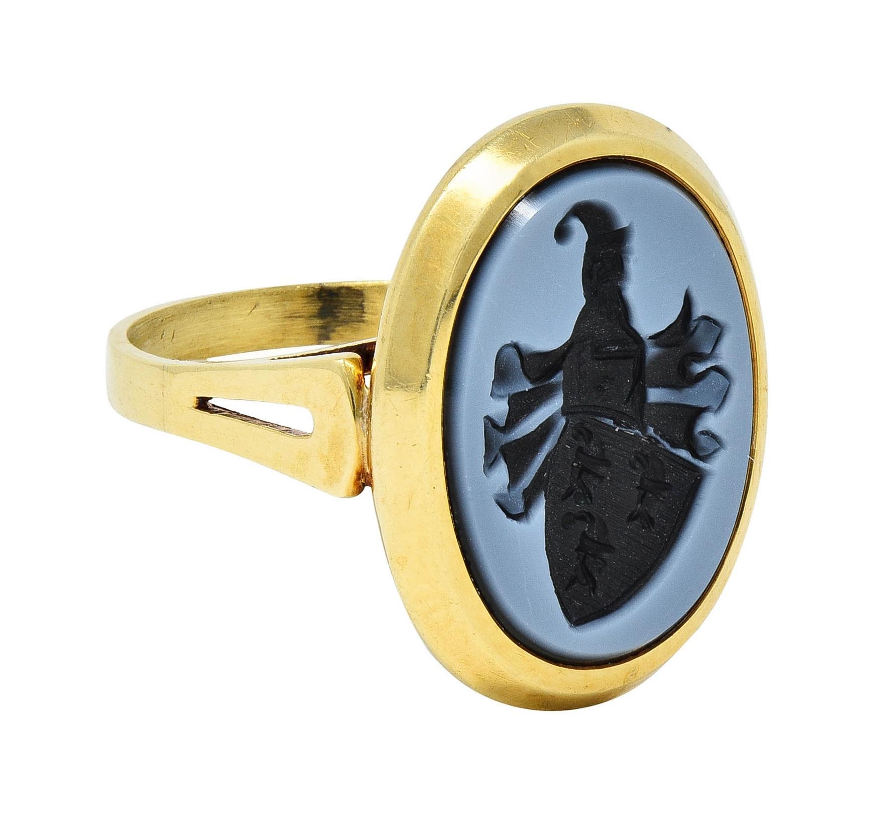 Centering an oval-shaped agate tablet measuring 15.0 x 10.5 mm
Banded opaque black and grayish-white - carved with intaglio
Depicting a heraldic coat of arms with banner and capped man
Set in a faceted bezel and flanked by pierced shoulders
Fanning