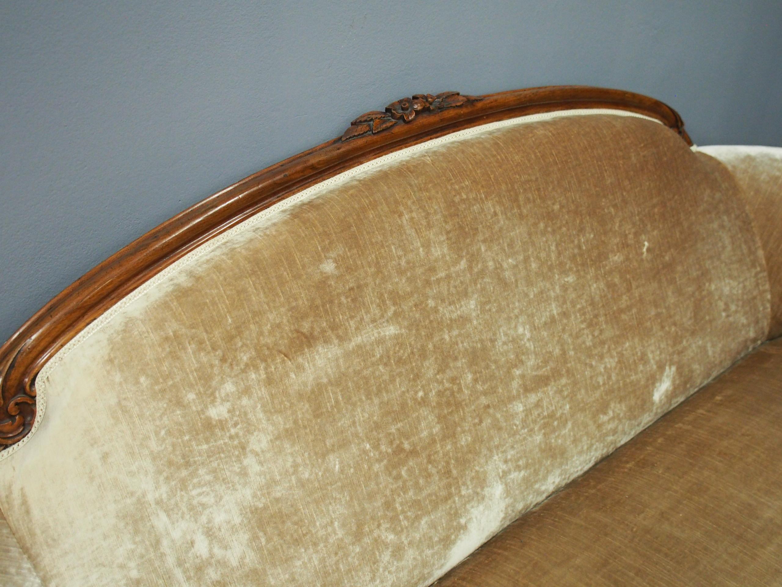 A carved walnut framed Victorian sofa, circa 1870. The solid walnut top rail at the back is carved with flowers and leaves, and it continues on to a beige-brown velour upholstery. The curved arm rests have a carved cabriole section at the front