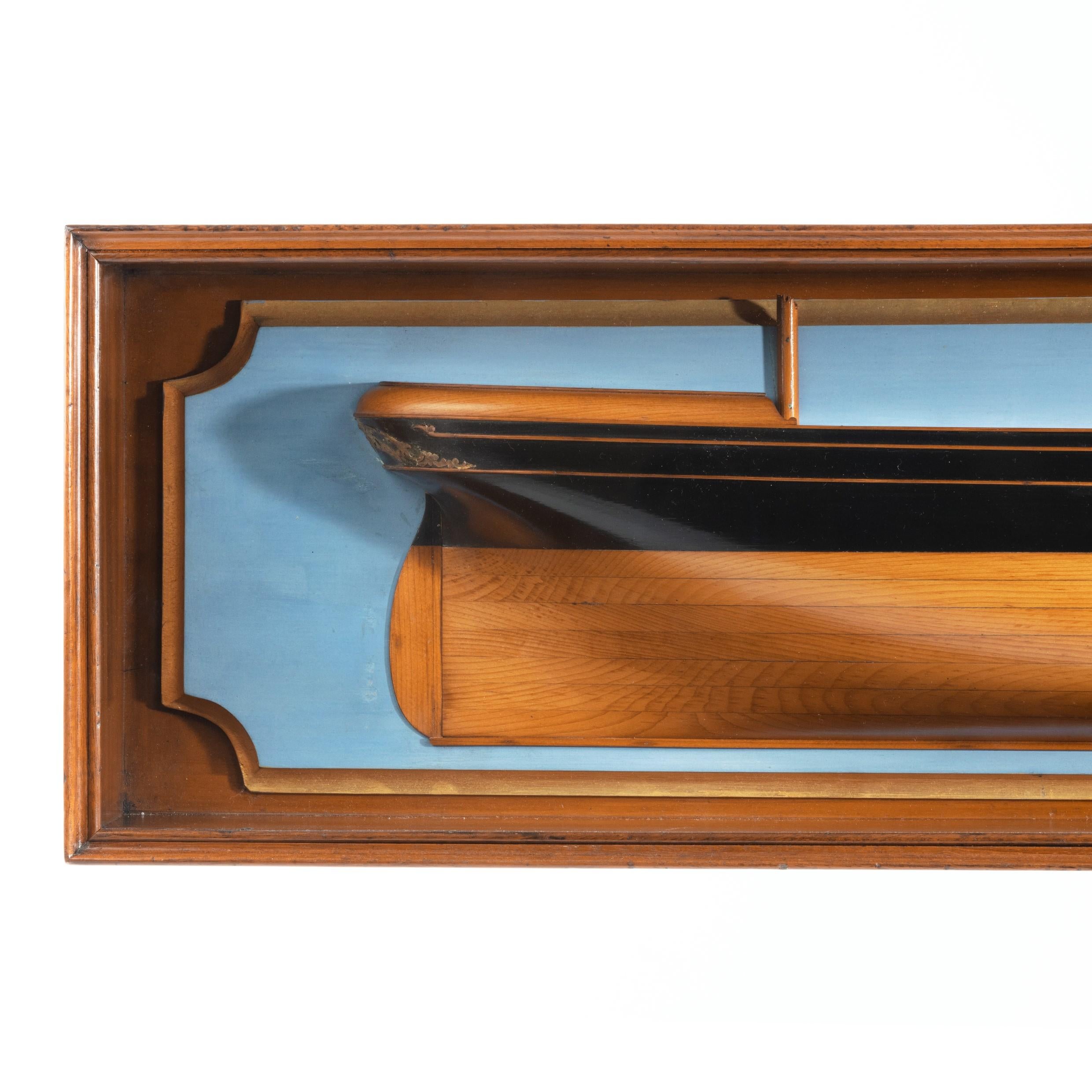 A Victorian cased half hull of a three masted sailing ship, shown with boxwood planking below the original black painted topsides, applied with gilt scrolling decoration to bow and stern, set on a shaped gilt and pale blue backboard, within a glazed