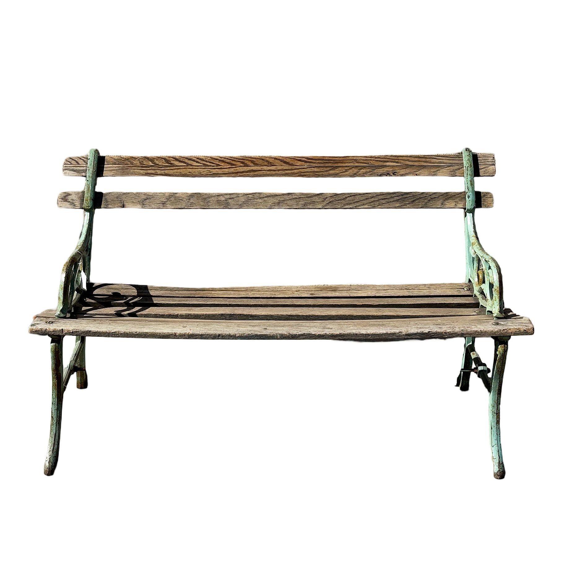 Encantador banco de jardín antiguo de hierro fundido y madera, con ornamentadas volutas laterales patinadas en verde pálido desgastado. El asiento y el respaldo de listones de roble muestran la edad y el carácter naturales, añadiendo un encanto