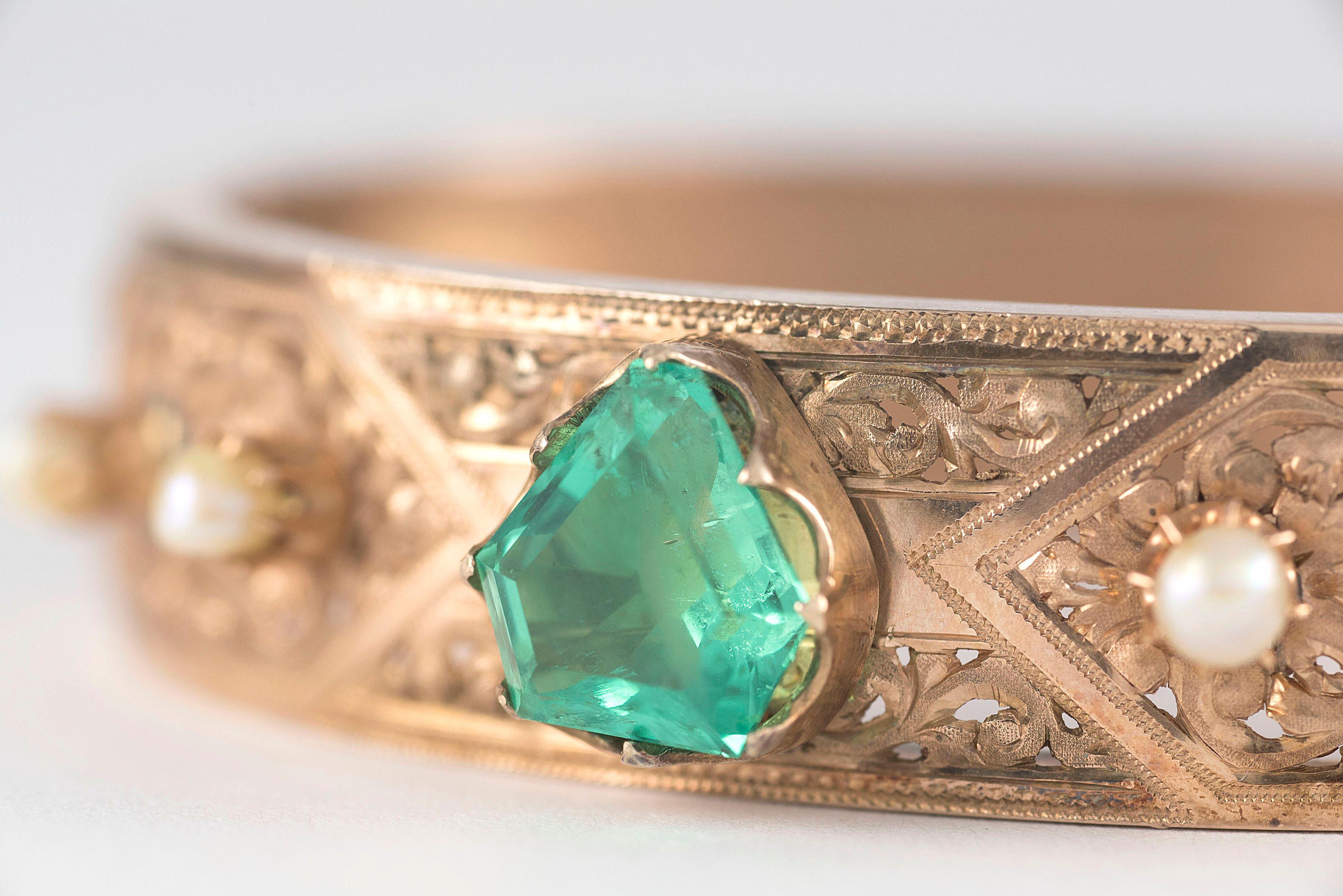 Crafted in 9kt rose gold, this antique Victorian bangle bracelet is designed around a natural unheated kite-cut Colombian emerald measuring 3.22 carats and surrounded by four natural white pearls and intricate decorative swirling piercing. 

