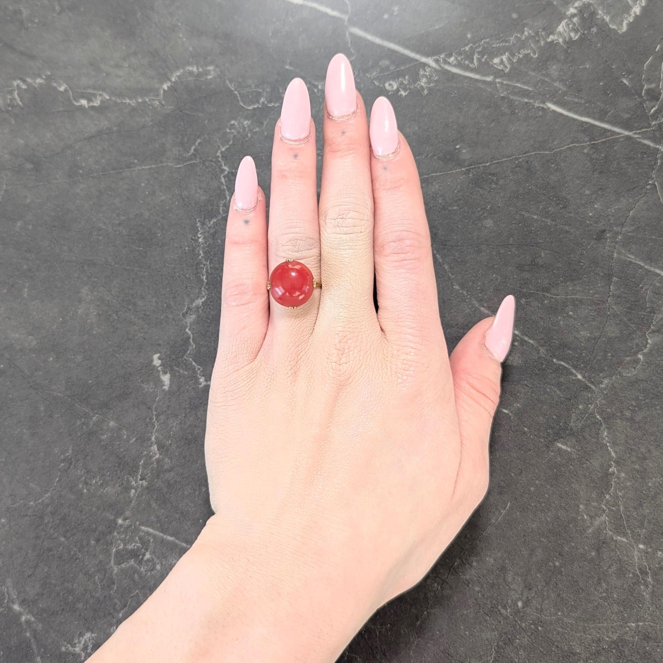 Centering a 14.5 mm round carved coral cabochon - opaque deep orangey red in color
Set with tri-split prongs in a scalloped basket pierced with a fanning motif
Flanked by tapered shoulders with grooved motifs
Accented by milgrain detailing
Stamped