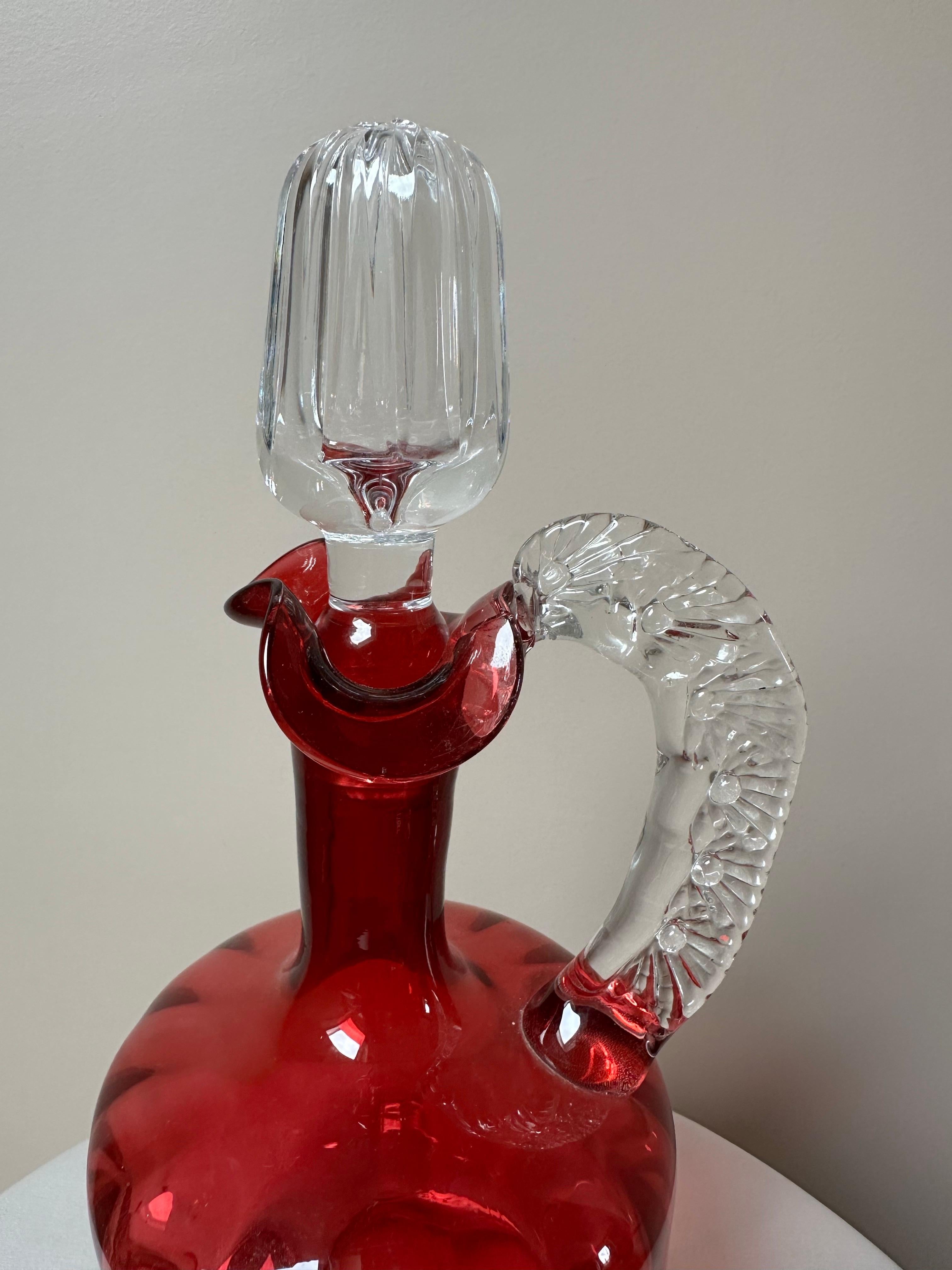 Vidrio soplado a mano, con un cuerpo de color rojo rubí intenso y detalles transparentes. El decantador tiene un cuerpo panelado con una base ancha que se estrecha en un cuello esbelto. El asa es de cristal transparente, texturizado con impresiones