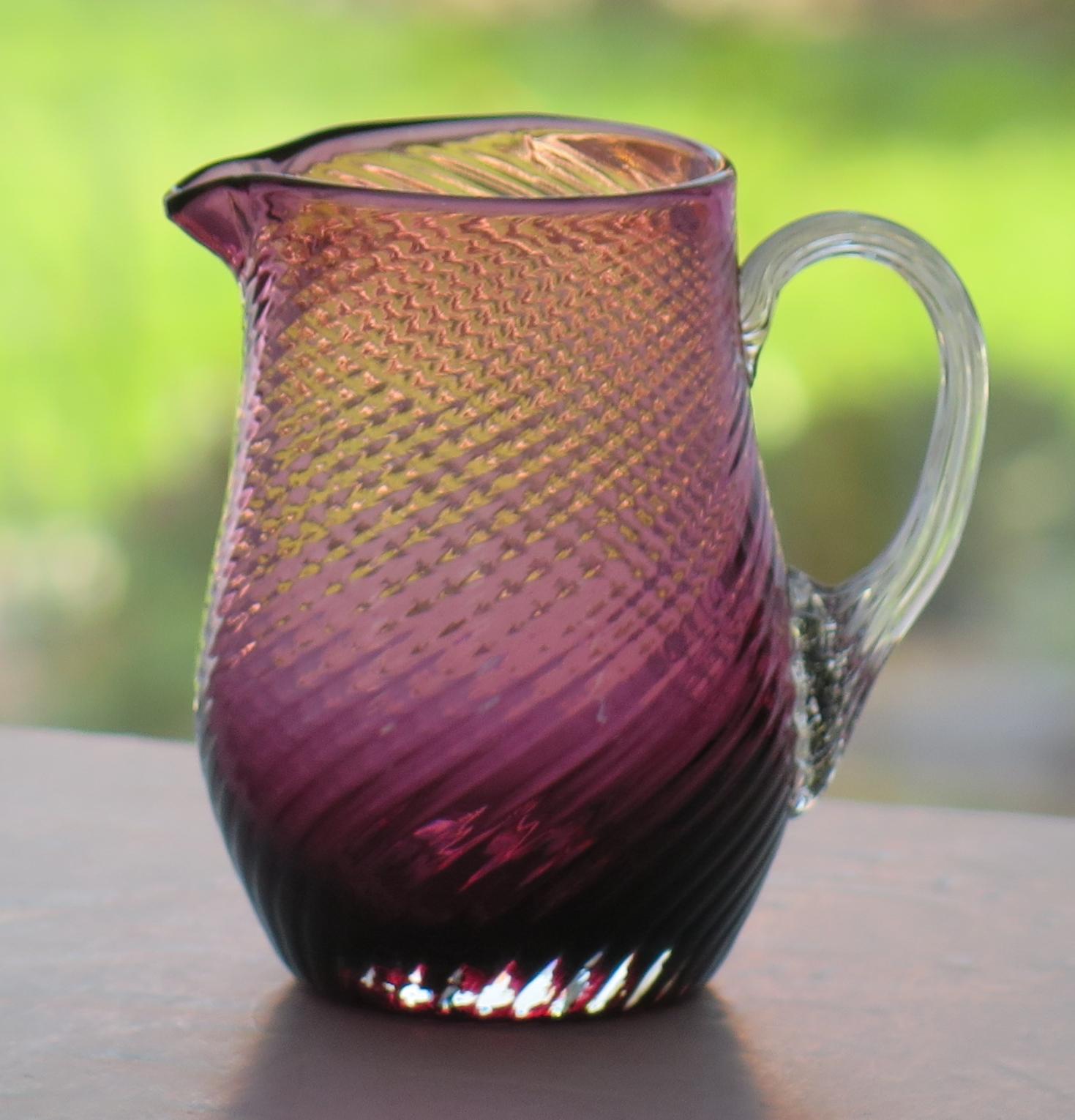 This is a good wrythen (spiral) moulded cranberry glass milk or cream jug, which we date to the mid-late 19th century of Victorian England, circa 1870.

The jug is hand made from cranberry glass with an applied vertically ribbed clear glass loop