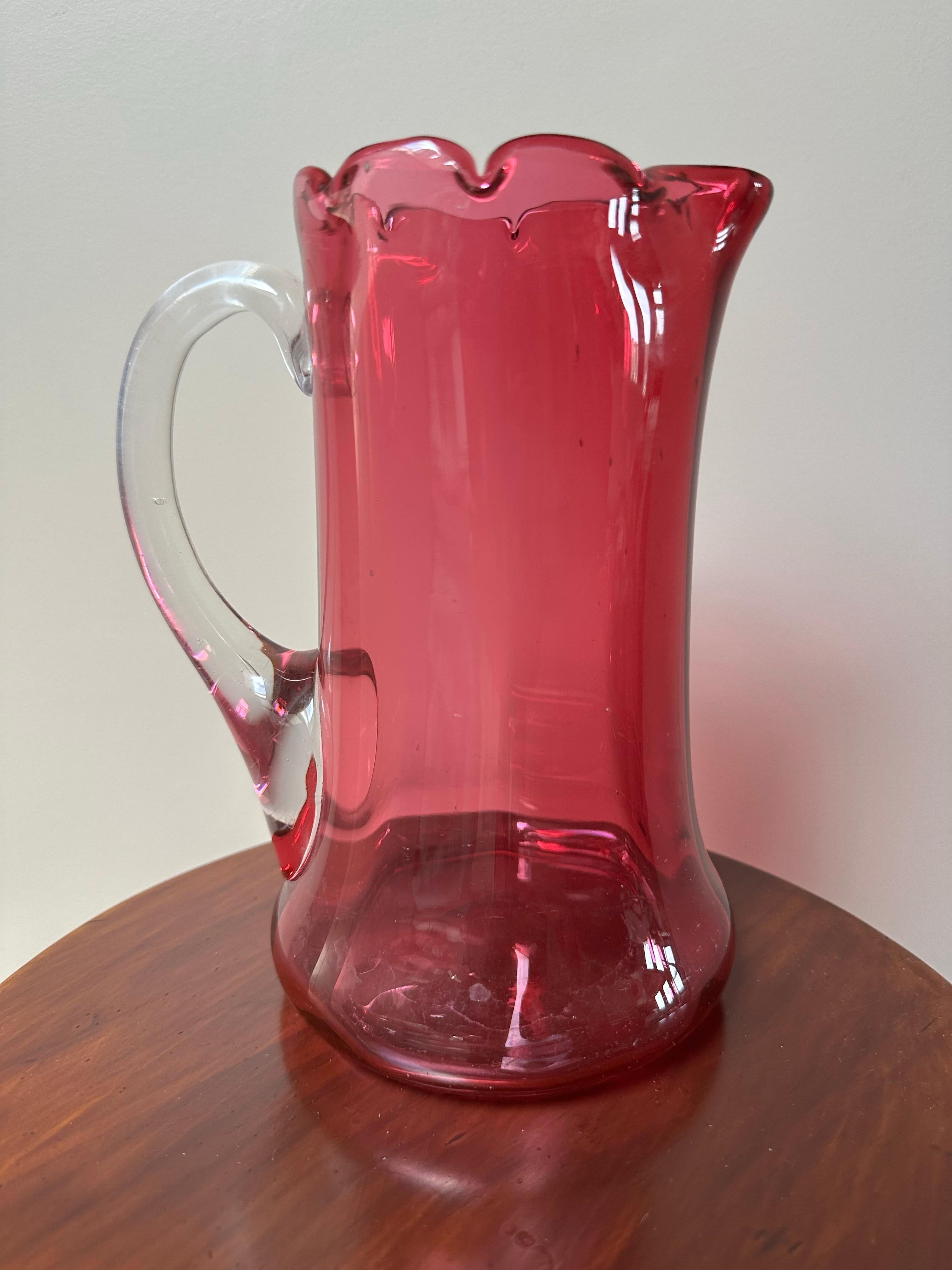 Questa elegante brocca per l'acqua è realizzata in vetro color mirtillo rosso, un materiale caratteristico ottenuto aggiungendo sali d'oro al vetro fuso. Risalente al 1900 circa, la brocca presenta un corpo aggraziato e svasato con una base ampia e