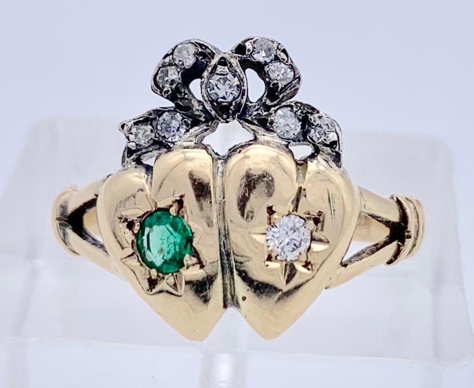 Two red gold hearts side by side, one is set with an emerald, the other with a diamond. They are joined by a silver  bow set with nine old cut diamonds. The ring is marked 15K on the inside of the shank. The ring is a typical example of sentimental