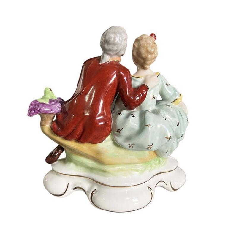 A ceramic figurine of a courting couple. This Victorian piece is created from a crisp white ceramic and features a man and woman perched upon a tree log. They lean into one another and share grapes. Each is hand-painted in an array of bright colors