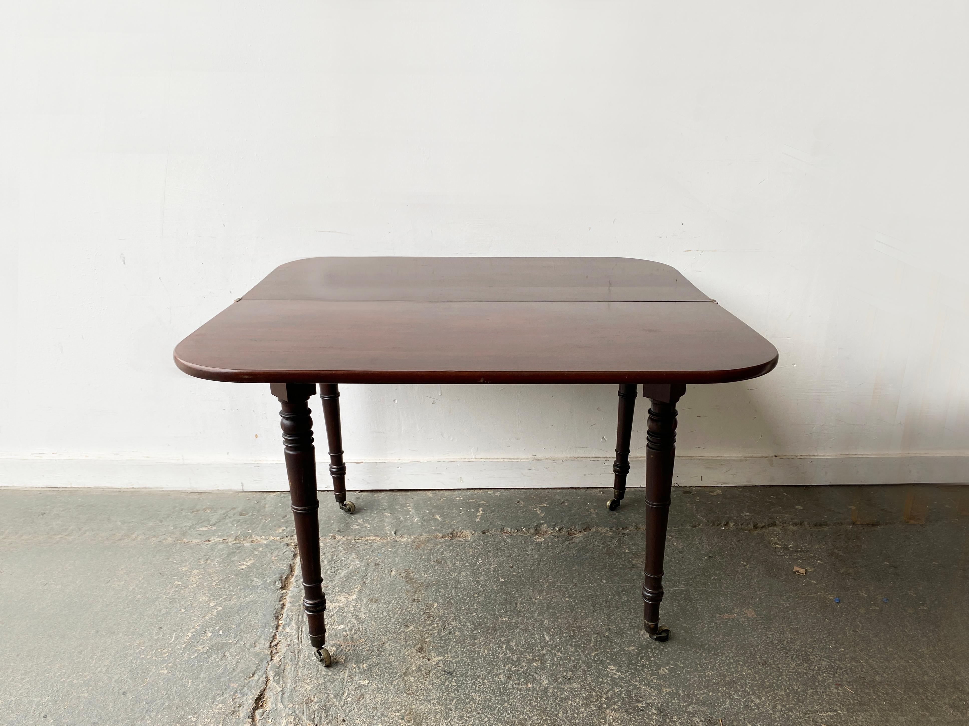 Table de salle à manger victorienne en acajou massif du milieu du 19e siècle. La table allongée a une forme carrée avec des coins arrondis, soutenue par quatre pieds tournés avec des roulettes d'origine en laiton et en céramique. 

La table est