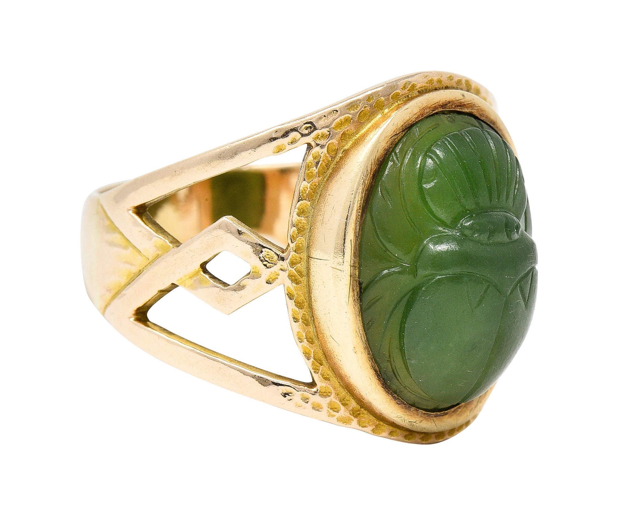 Centering an oval shaped nephrite cabochon carved to depict a scarab beetle. Measuring 13.5 x 10.5 mm - translucent medium to dark green. Bezel set with a hammer texture surround. Flanked by pierced 'W' motif shoulders. Tested as 14 karat gold.