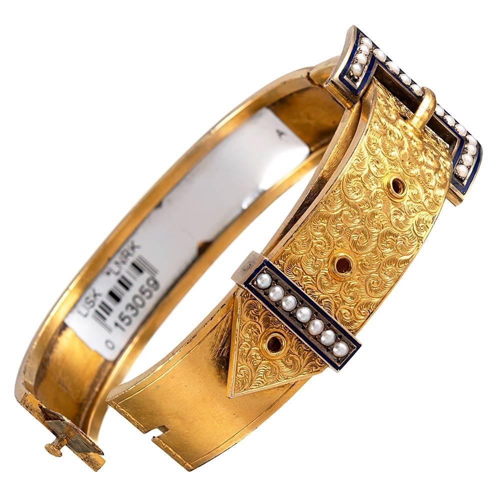 18 karat yellow gold bangle, extensively decorated with scrolling hand engraving and accented with royal blue enamel with seed pearls. The interior diameter measures 2 1/4 by 1 7/8 inches. This is a lovely example of antique accessory and can be