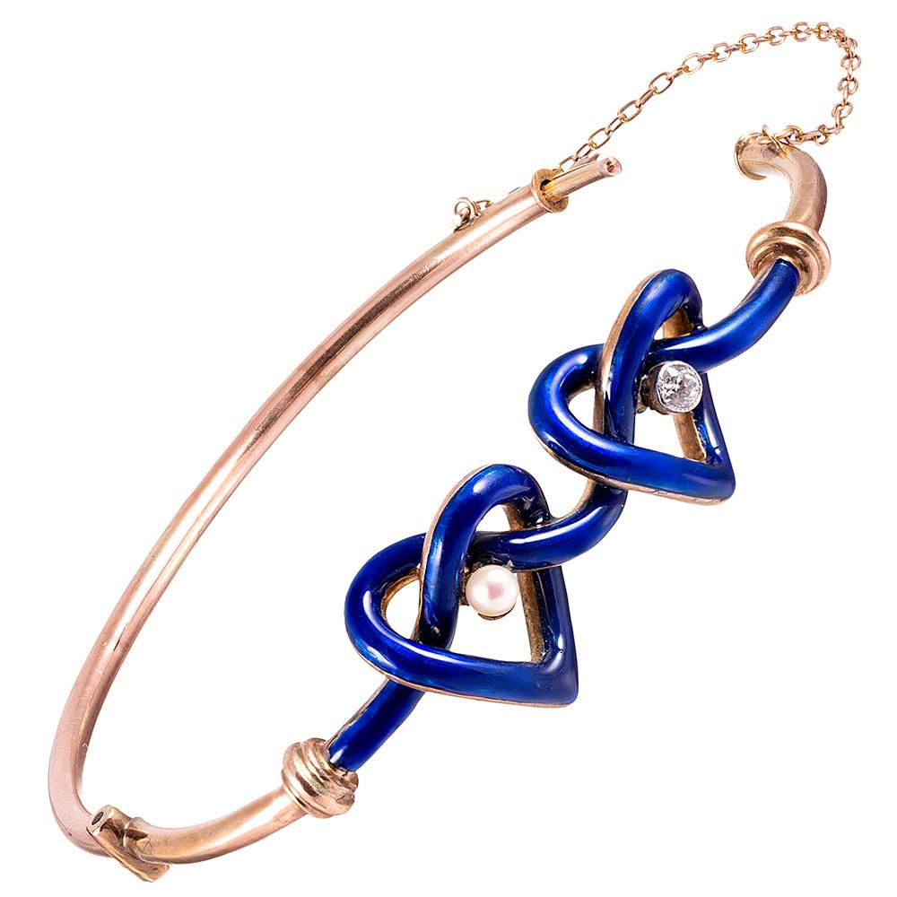 Made in England, this late Victorian bracelet is abundant with charm and romanticism. 9 carat yellow gold and intense blue enamel forms a double heart-shaped “lover’s knot”, one set in the center with a diamond and the other a pearl. Finished with a