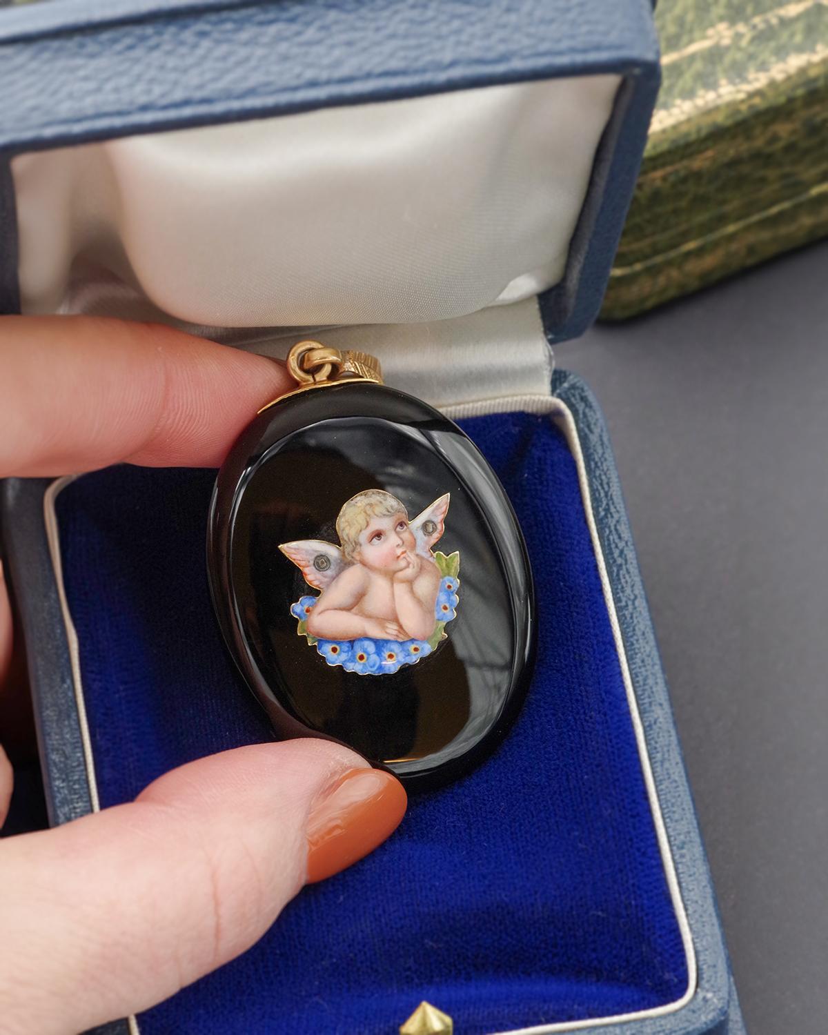 Onyx angel locket pendant in 18 karat gold. This antique locket features an oval onyx angel depicting a pensive cherub resting on a bed of forget-me-nots. Its blond hair and rosy complexion contrast with the deep black of the stone. The back of the