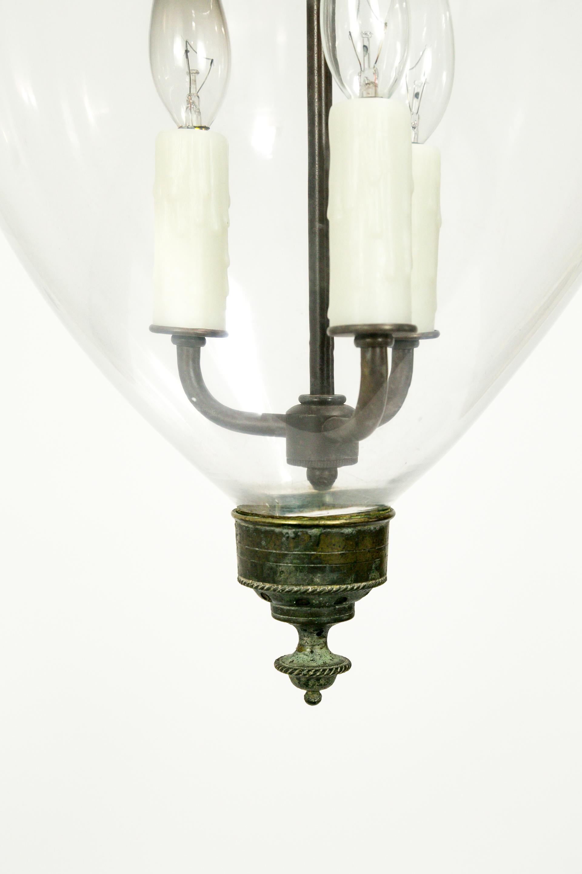 An elegant English bell jar lantern with refined details on the brass hardware: a ribbed glass holder with bird head chain hooks, and a rope-trimmed finial. The brass has a beautiful, aged patina. Handblown jar and smoke bell. 3-light candelabra