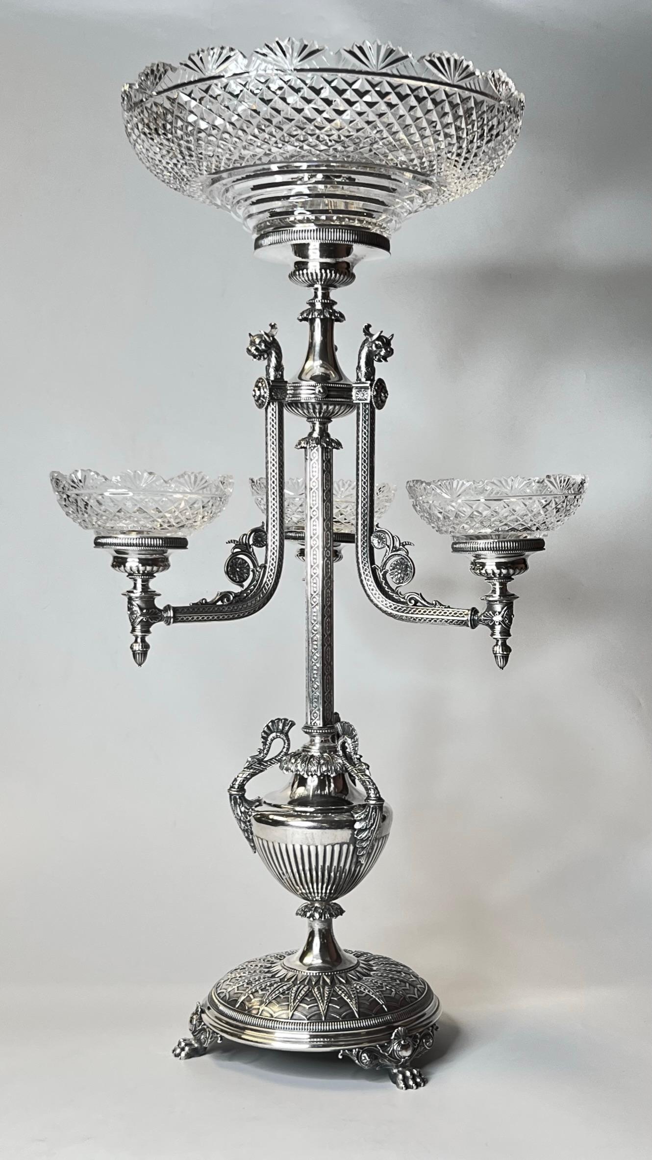 Notre épergne en argent sterling de la firme Manoah Rhodes and Sons de Bradford, vers 1888-1890, présente de merveilleux motifs néoclassiques, notamment des animaux à cornes et des oiseaux exotiques, un plat principal en verre taillé et trois petits