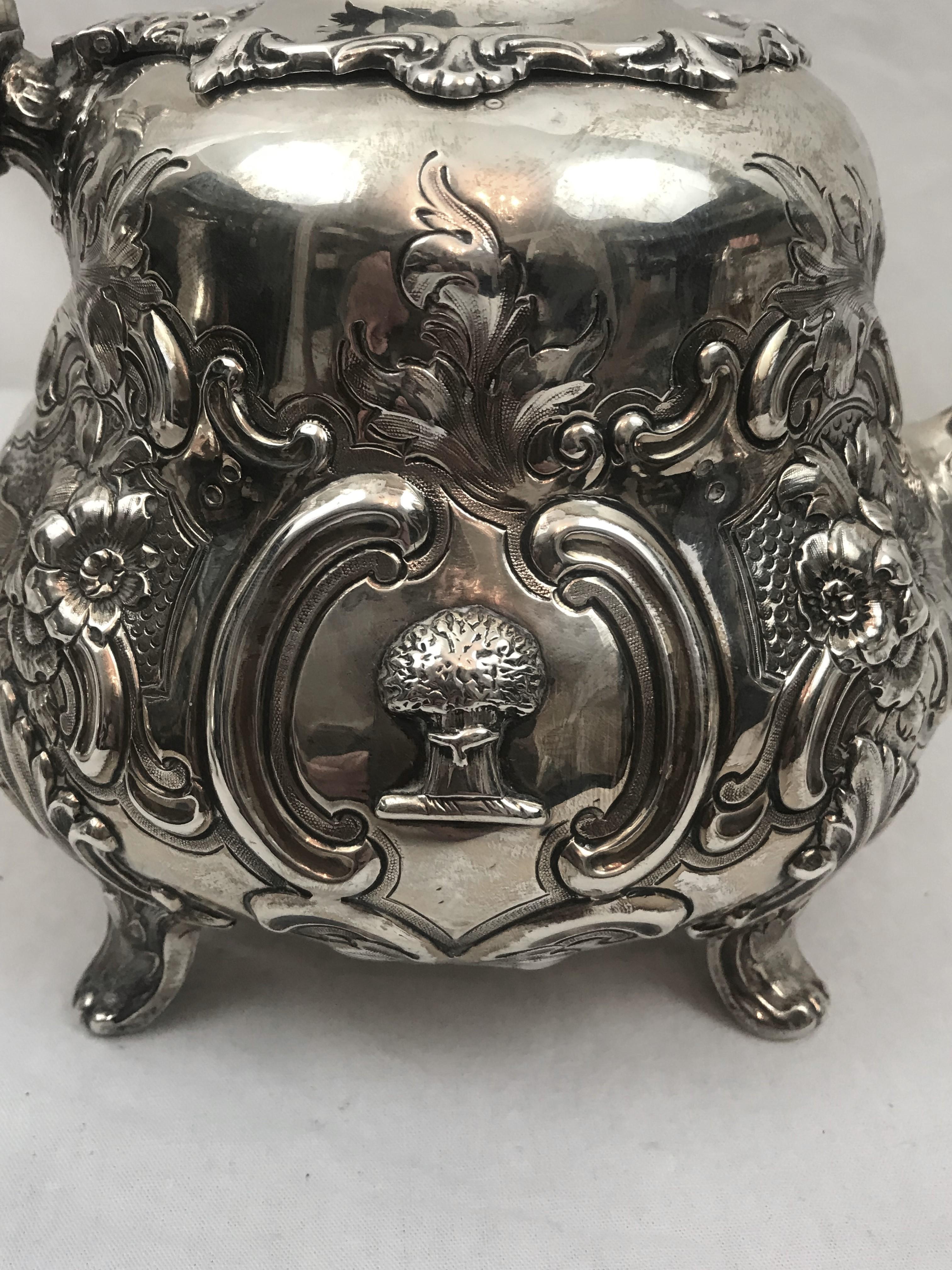 English sterling silver footed teapot. 
Date mark London 1855. Maker's mark William Hunter.
 
Heavily repoussed flowers and scrolling leaves. Flower finial. 
Unusual applied family crest of a tree or sheath of wheat.
Bone insulators.
