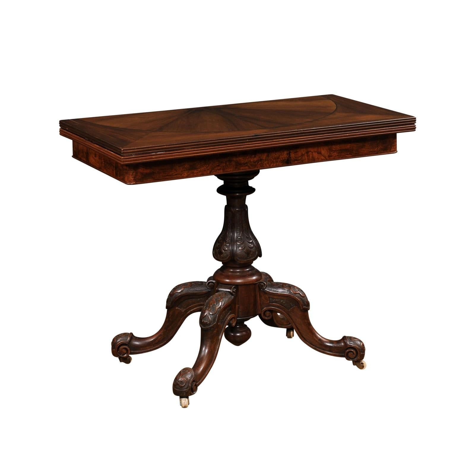 An English Victorian period fold-over walnut mahogany game table from the 19th century with bookmatched top and carved base on casters. Step into the elegance of the Victorian era with this exquisite English fold-over mahogany game table from the