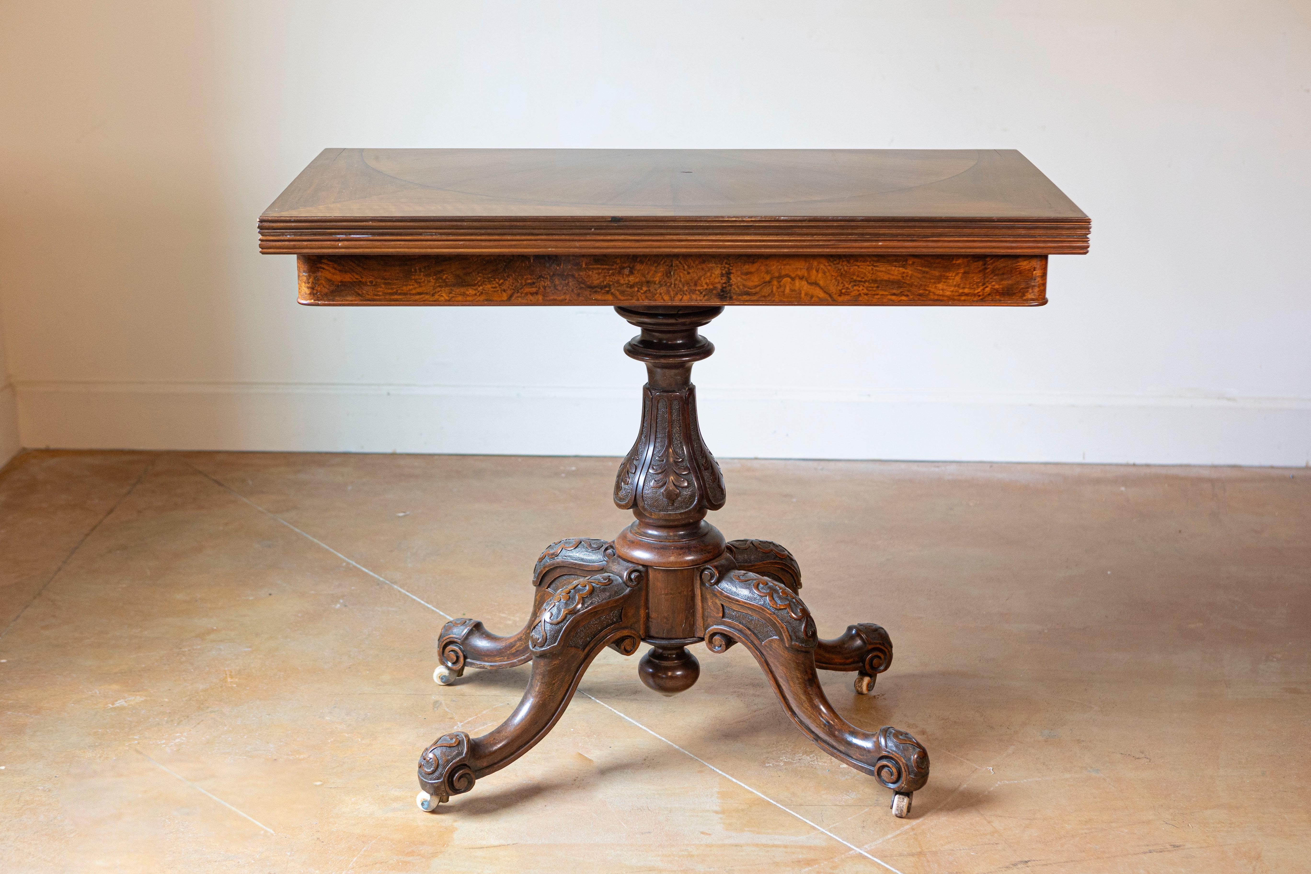 An English Victorian period fold-over walnut mahogany game table from the 19th century with bookmatched top and carved base on casters. Step into the elegance of the Victorian era with this exquisite English fold-over mahogany game table from the