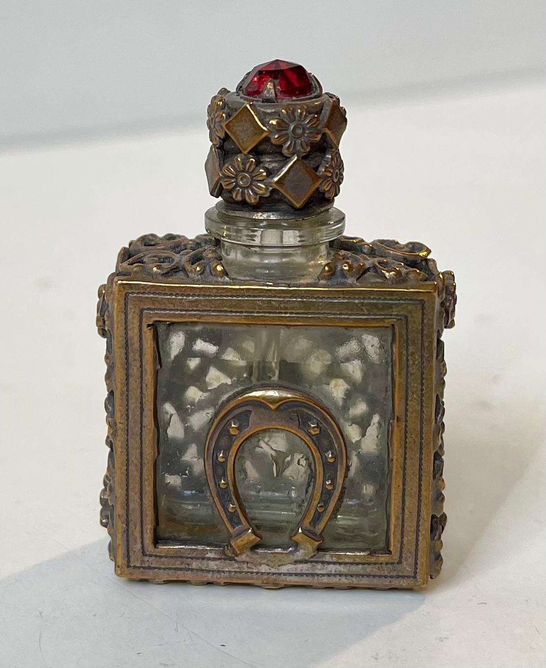 Miniature perfume or scent bottle in crystal and ornate brass. Made in Europe, probably England circa 1850-1890. It features a horseshoe and it has a faceted garnet or red piece of glass to its lid. Measurements: 4.5x3x2 cm.