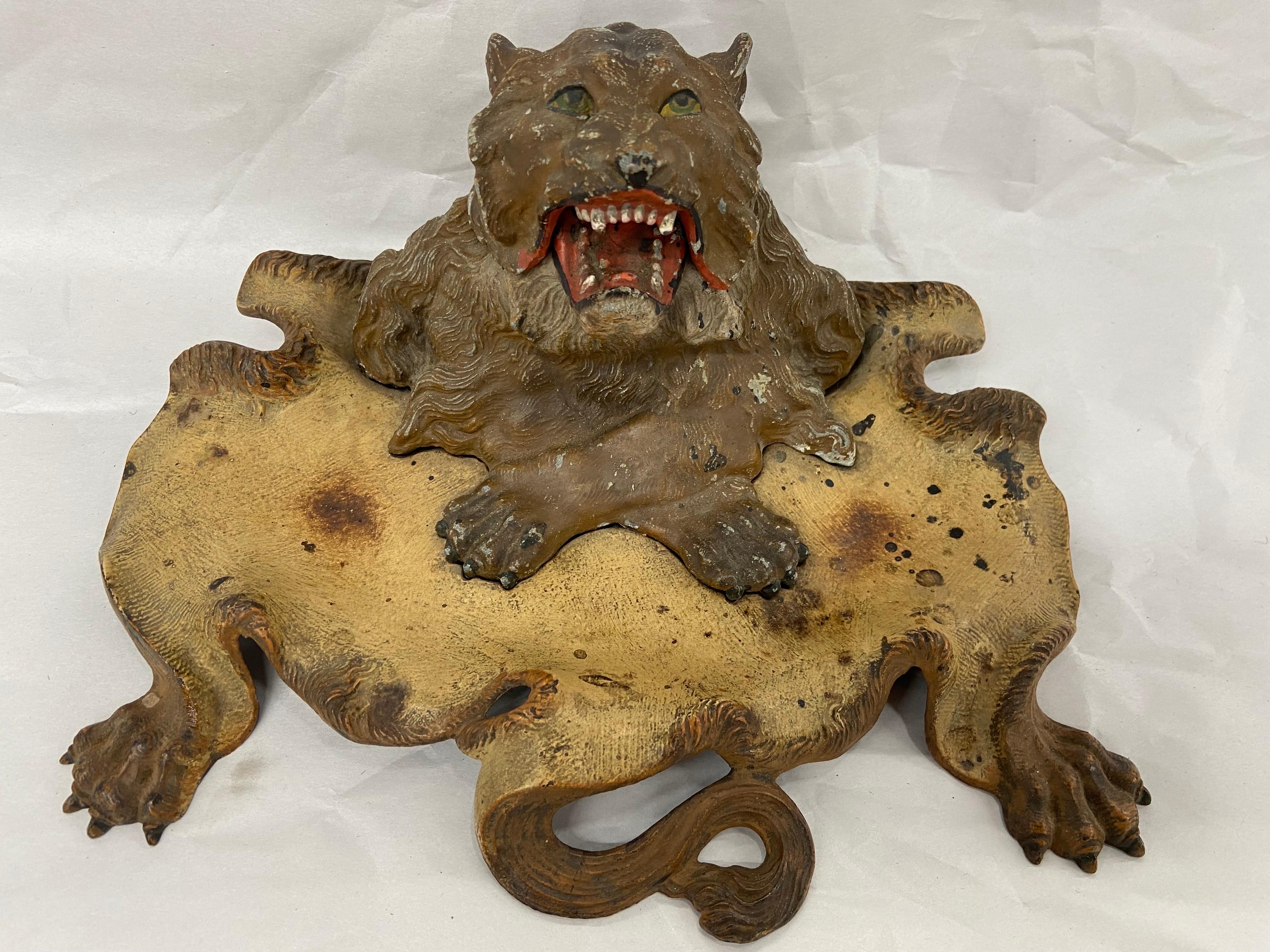 A Victorian Era, circa 1900, antique cold painted spelter (smelted and alloyed zinc) figural inkwell depicting a roaring lion with front paws crossed and a lion skin rug that serves as a pen rest or even a vide poche. The base / lion skin rug