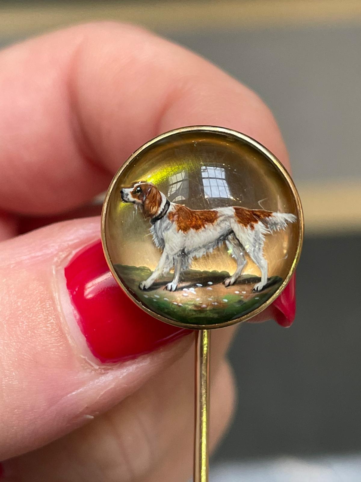A Victorian Essex Crystal spaniel dog stickpin 18 karat yellow gold.

This charming pin features a domed crystal with an exquisite collie dog scene. The white dog displays brown patches and wears a mahogany coloured collar. It stands on green