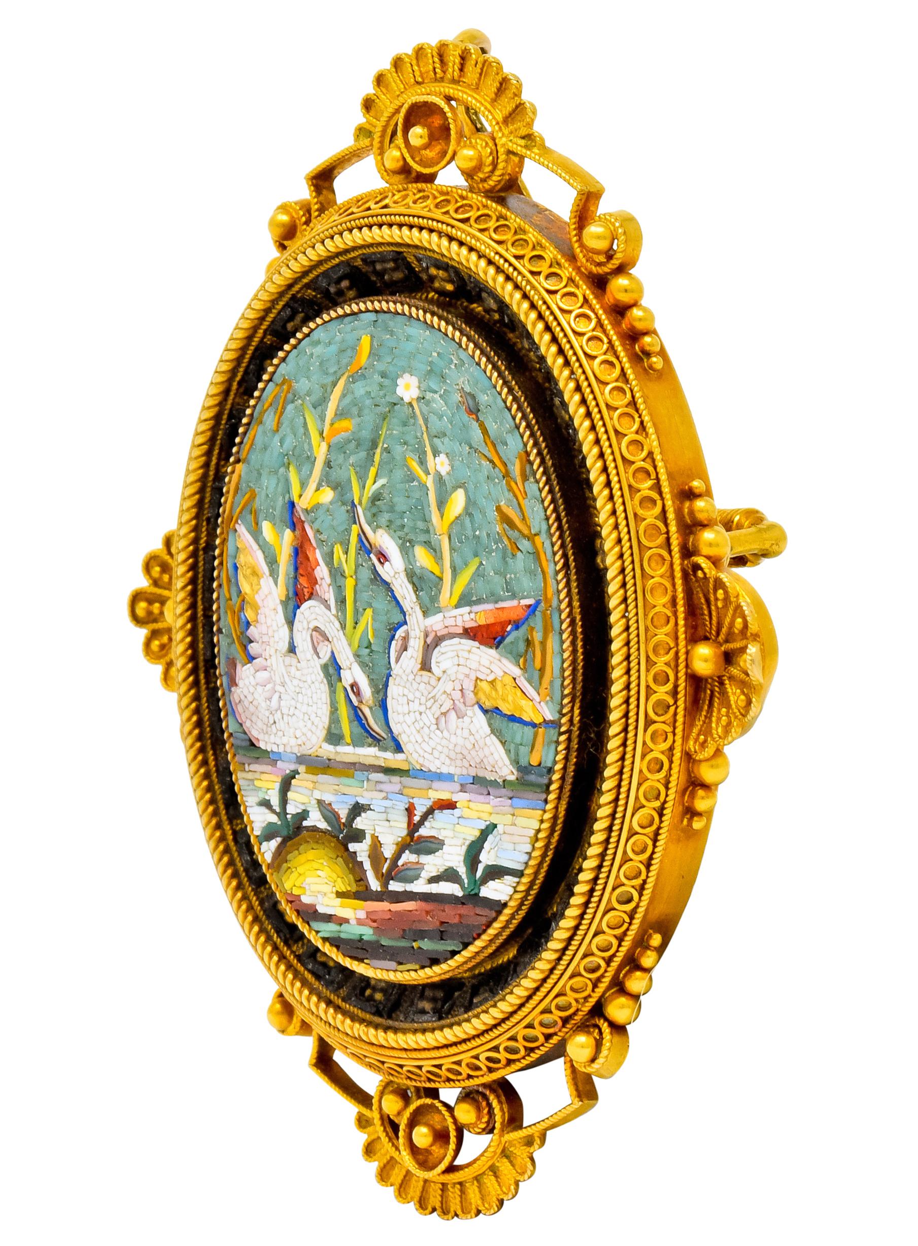 Pendant brooch designed as an ornate frame with scalloped accents at each cardinal point, a circle motif surround, and gold bead detail throughout

Centering an oval depiction of two playful swans floating in water amongst flowers and foliate