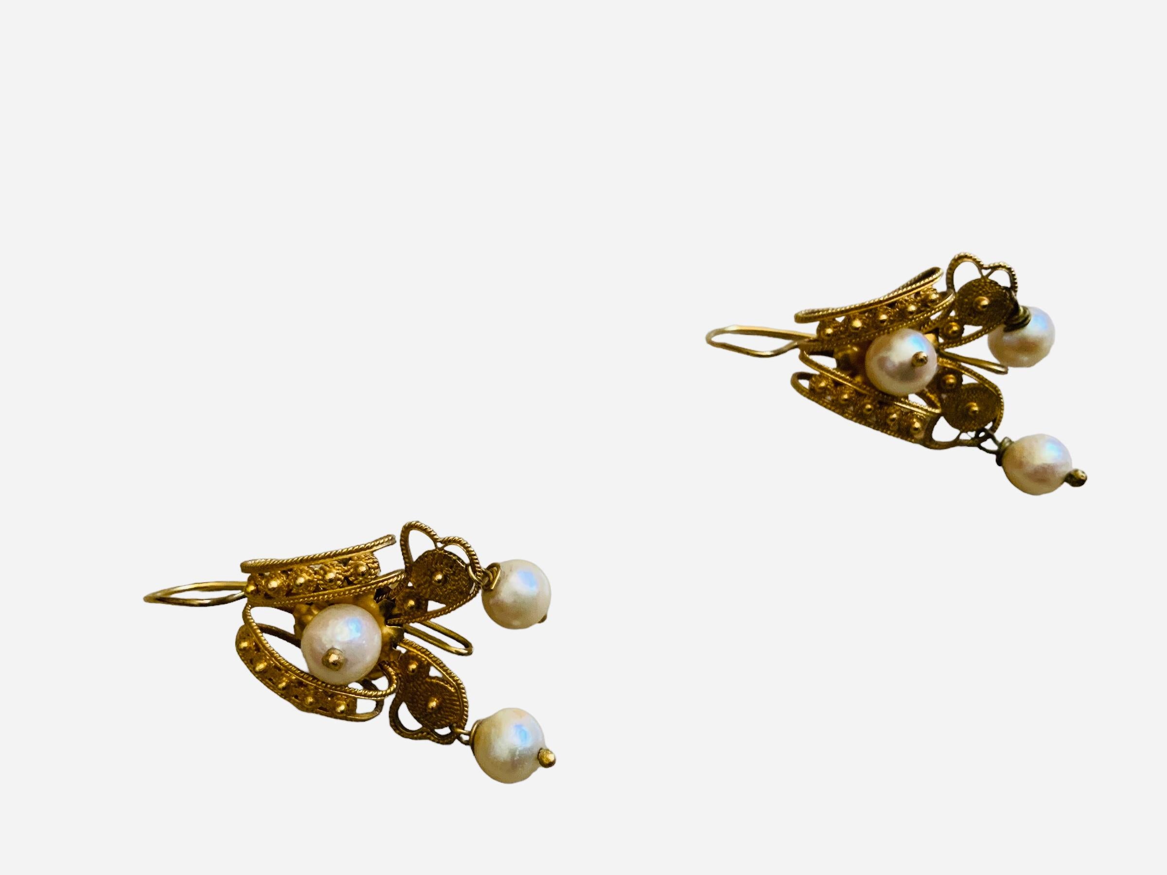 This is a 18K gold and pearls pair of Victorian Etruscan Style earrings. It depicts a pair of earrings adorned with delicate cannetille work and embellished with three pearls- two of them are dangling coquettishly. They use wire hooks.