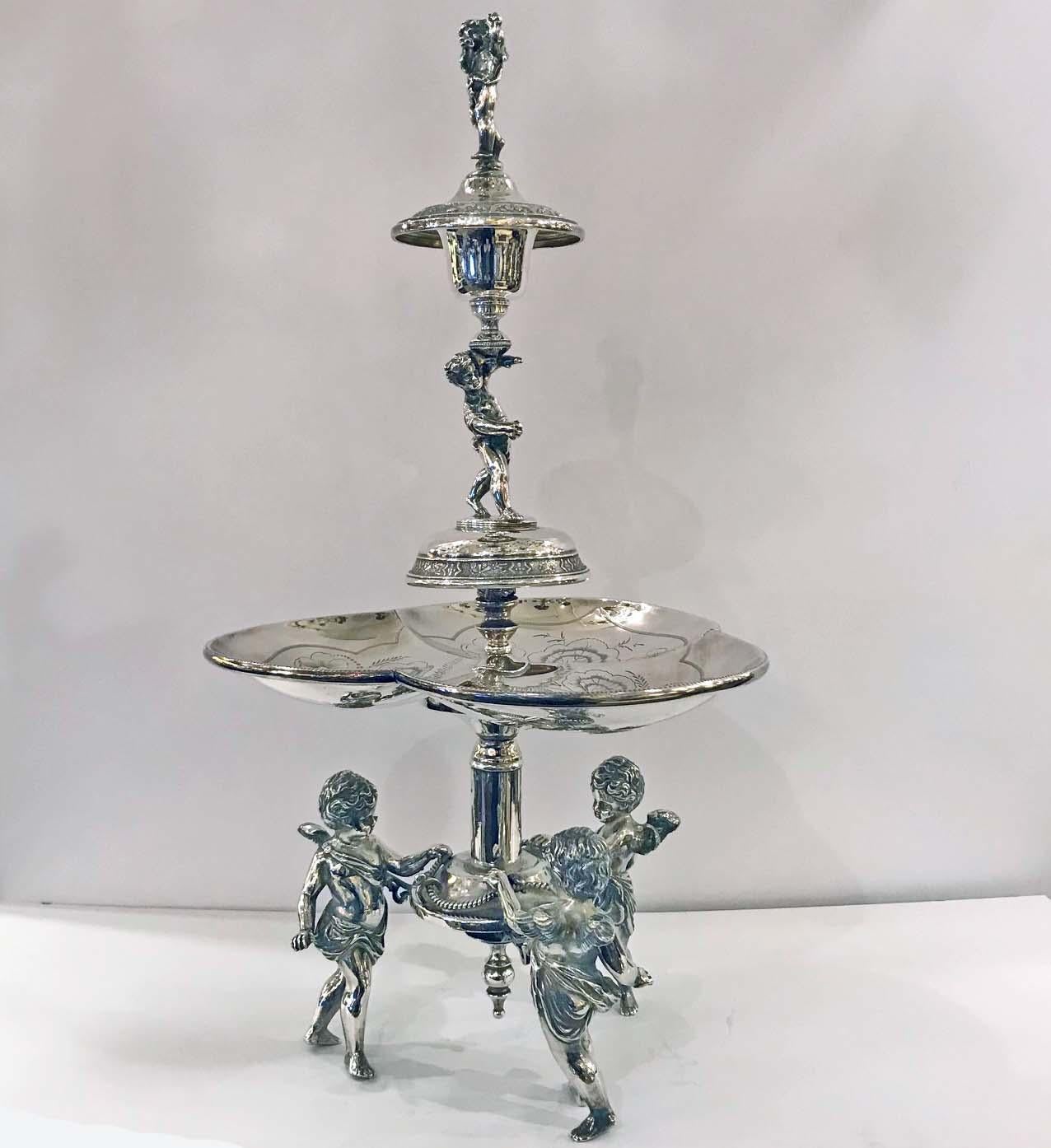 This exuberant centerpiece is a playful addition to your dinner table. The central trilobed dish is hand-engraved with flowers and supported by three frolicing cherubs. The elaborate finial is surmounted by a cherub sounding a blast on a horn: he is