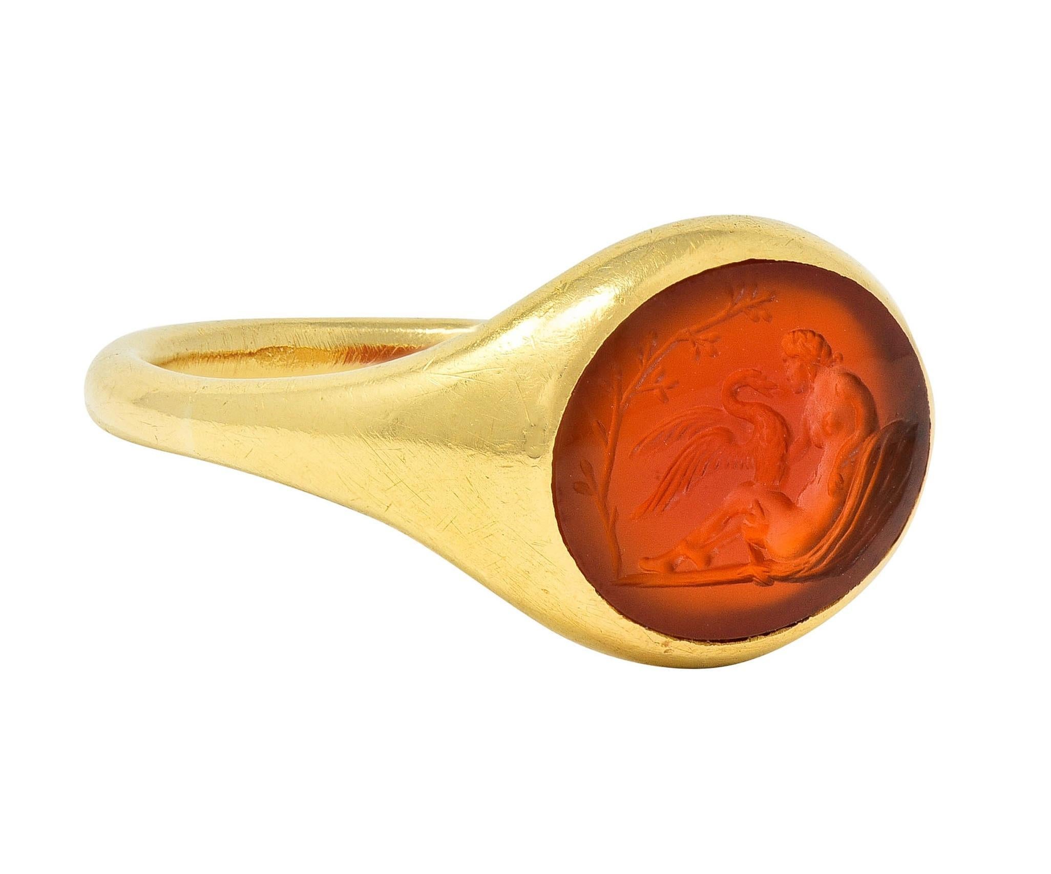 Centering an oval-shaped carnelian tablet carved with intaglio depicting a woman and swan
Based on the Greek myth of the figure Leda and The Swan (Zeus) 
Translucent reddish-orange and measuring 10.0 x 13.0 mm 
Flush set with a contoured gold