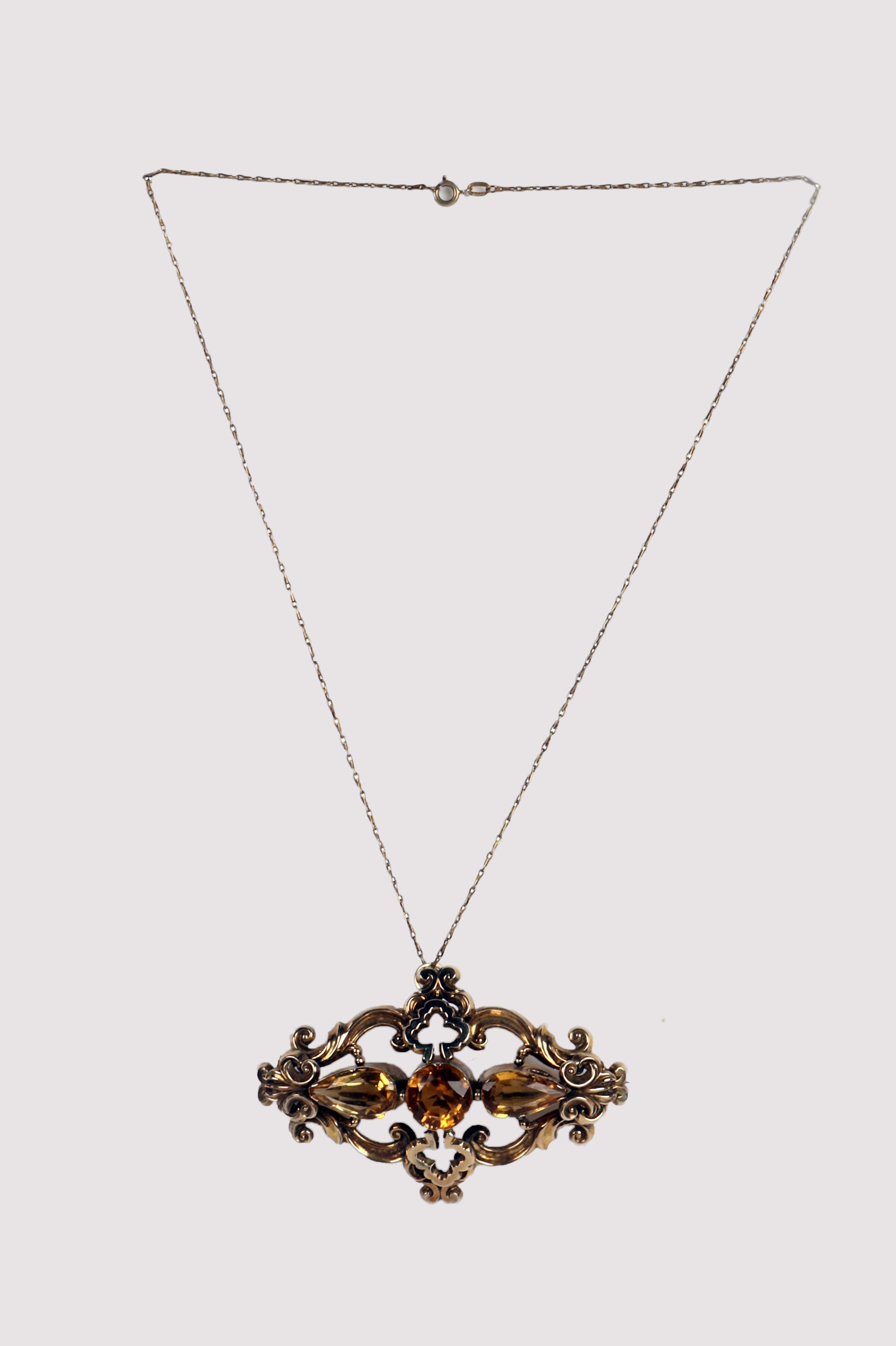 A thin gold necklace with elongated links and a circular lobster clasp holds a pendant brooch with a central body and oblong sides. Made of 14 Kt gold with box-shaped foil, it presents the joining elements of the frame designed with curvilinear