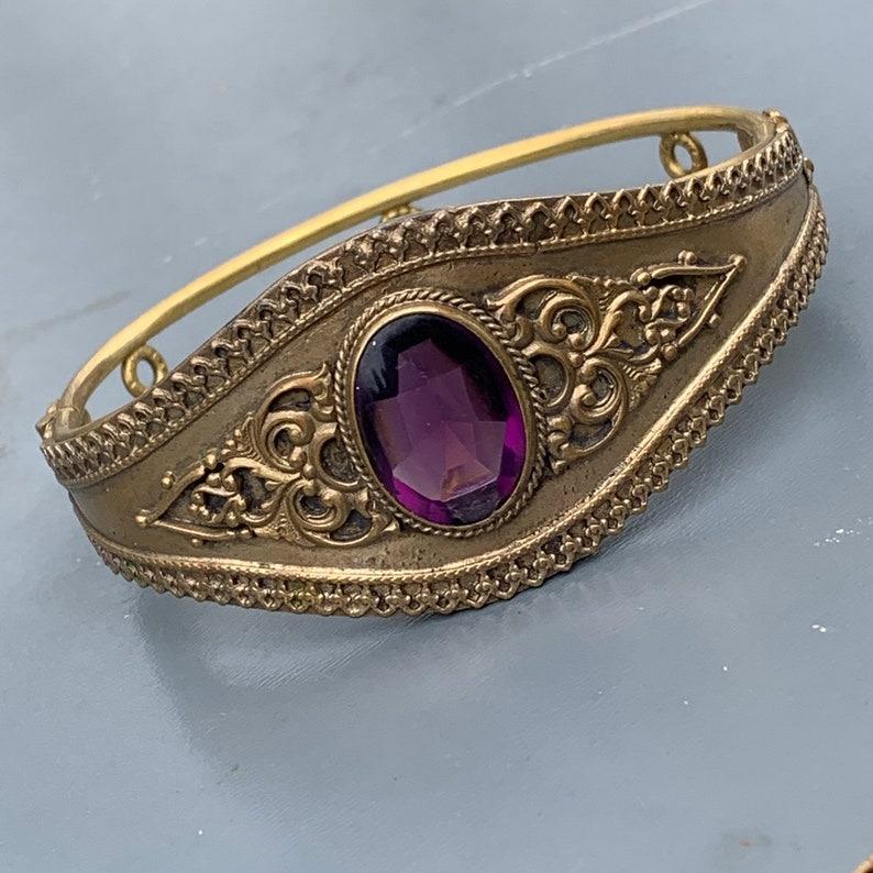 Le bracelet victorien antique est une pièce intemporelle, dotée d'un design à charnière avec un fermoir caché qui s'ouvre à l'aide d'un bouton poussoir discret. Réalisé en métal doré, ce bracelet est orné de pierres en pâte de verre améthyste, la