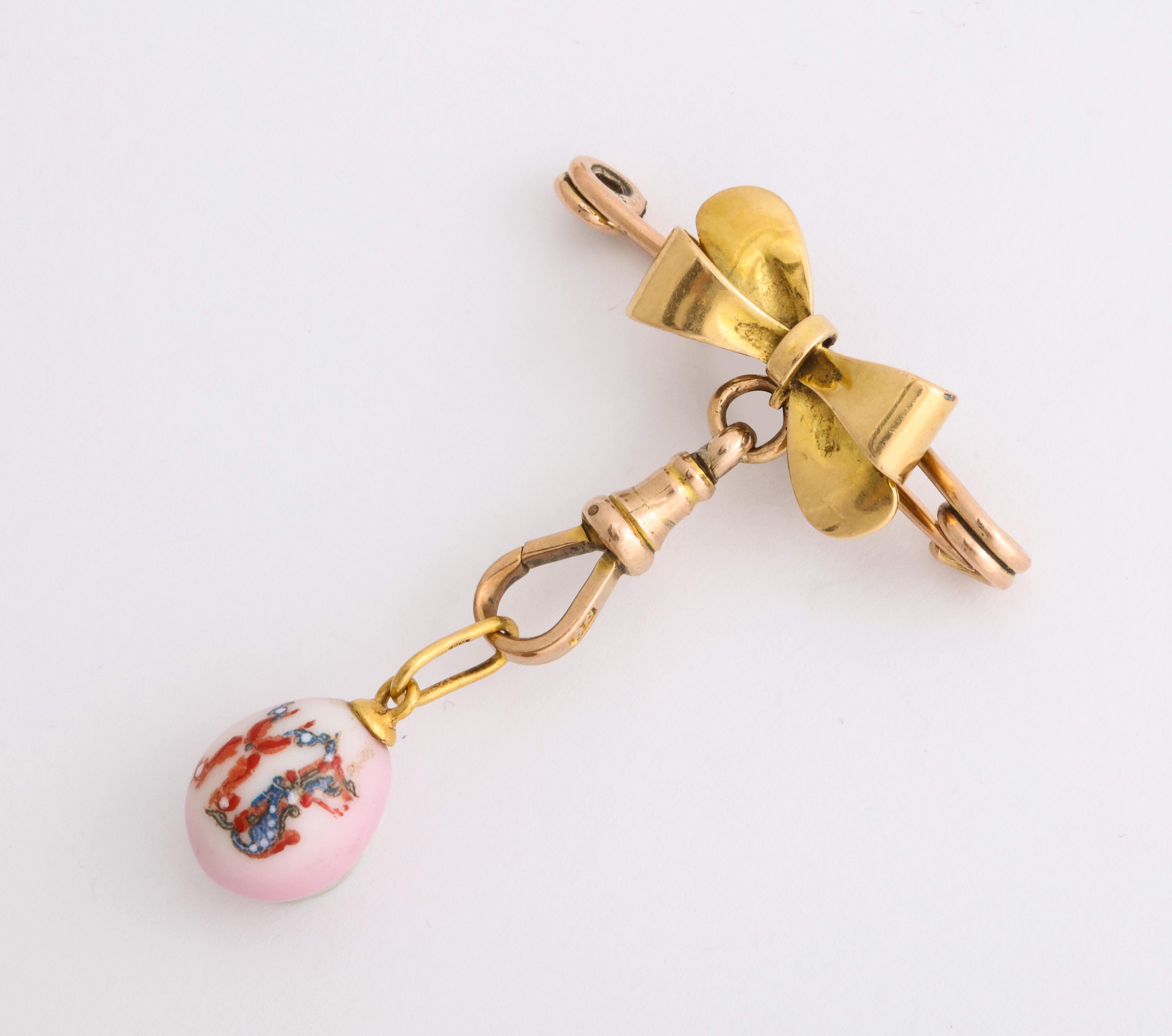 English Victorian pin designed as an 18k yellow gold bow on a 14k rose gold pin, suspending a Russian hand-painted porcelain gilded egg pendant, inscribed XB for Xristos Voskrese, a Russian Easter proclamation on pink ground. 

The pin circa 1890,