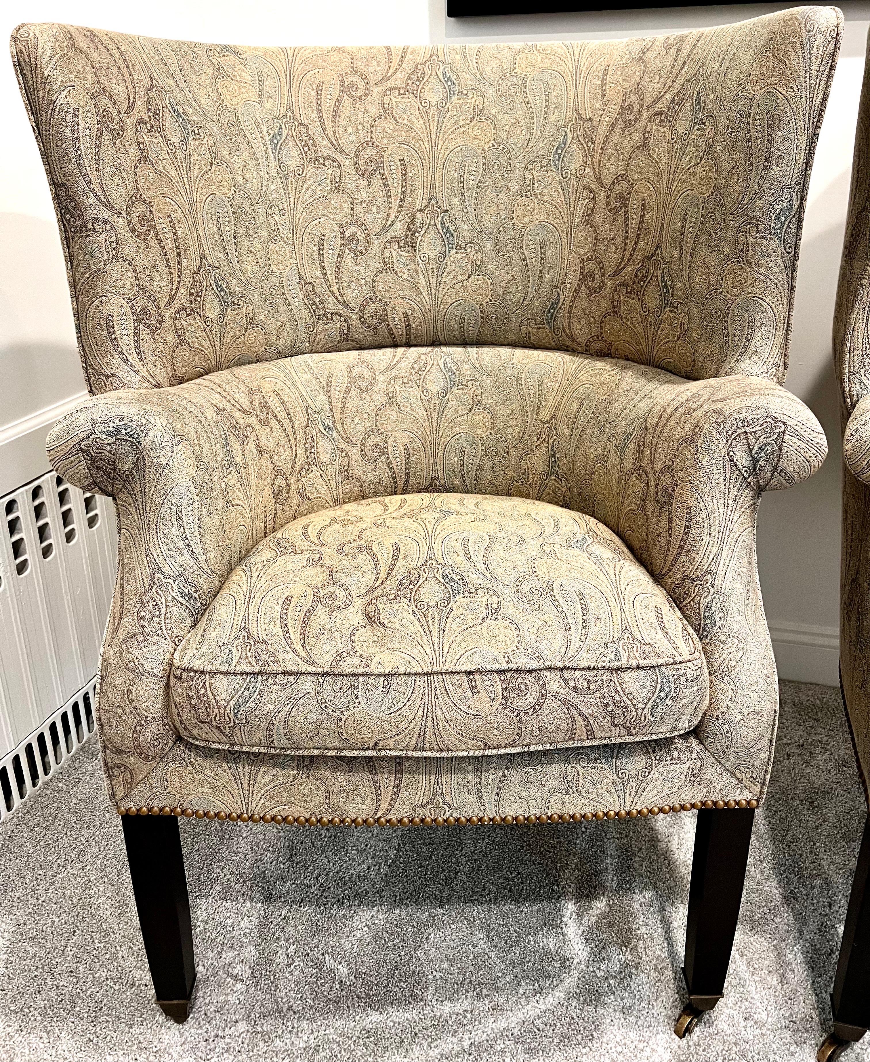 Beautiful pair of Wainscott wingback chairs by Victoria Hagan upholstered in paisley print in muted tan, olive and gray. They feature a wider wing profile and brass nailheads all around. Down seat cushion adds extra comfort.