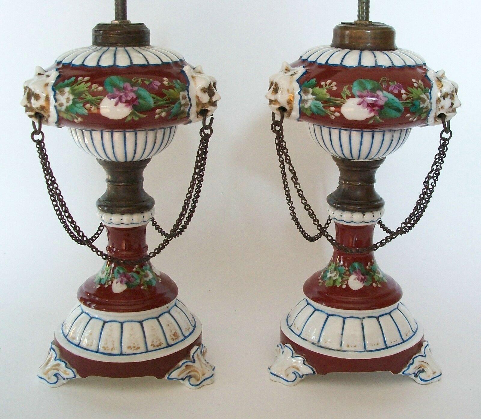 Fine pair of Victorian hand painted and gilded ceramic oil lamps - each featuring upper and lower floral bands - the top band separated by three gilded lion head masks with original bronze rings and chain swags - set against a sold burgundy
