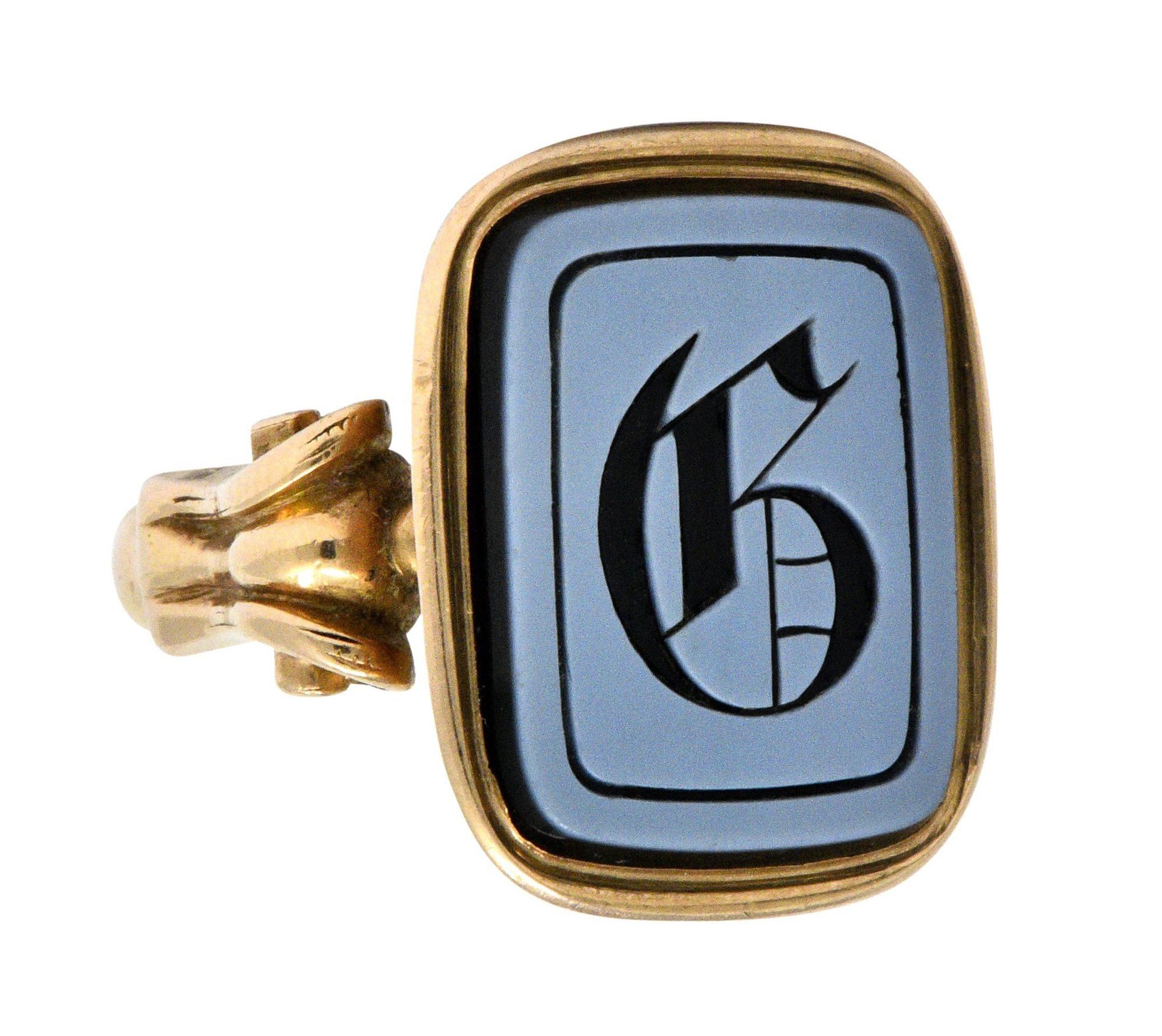 Centering an intaglio carved reverse uppercase G in banded only, measuring approximately 16.5 x 12.5 mm

With a white surface, black back, and a deeply engraved stylized black letter

Bezel set in a polished gold surround with millegrain detailing,