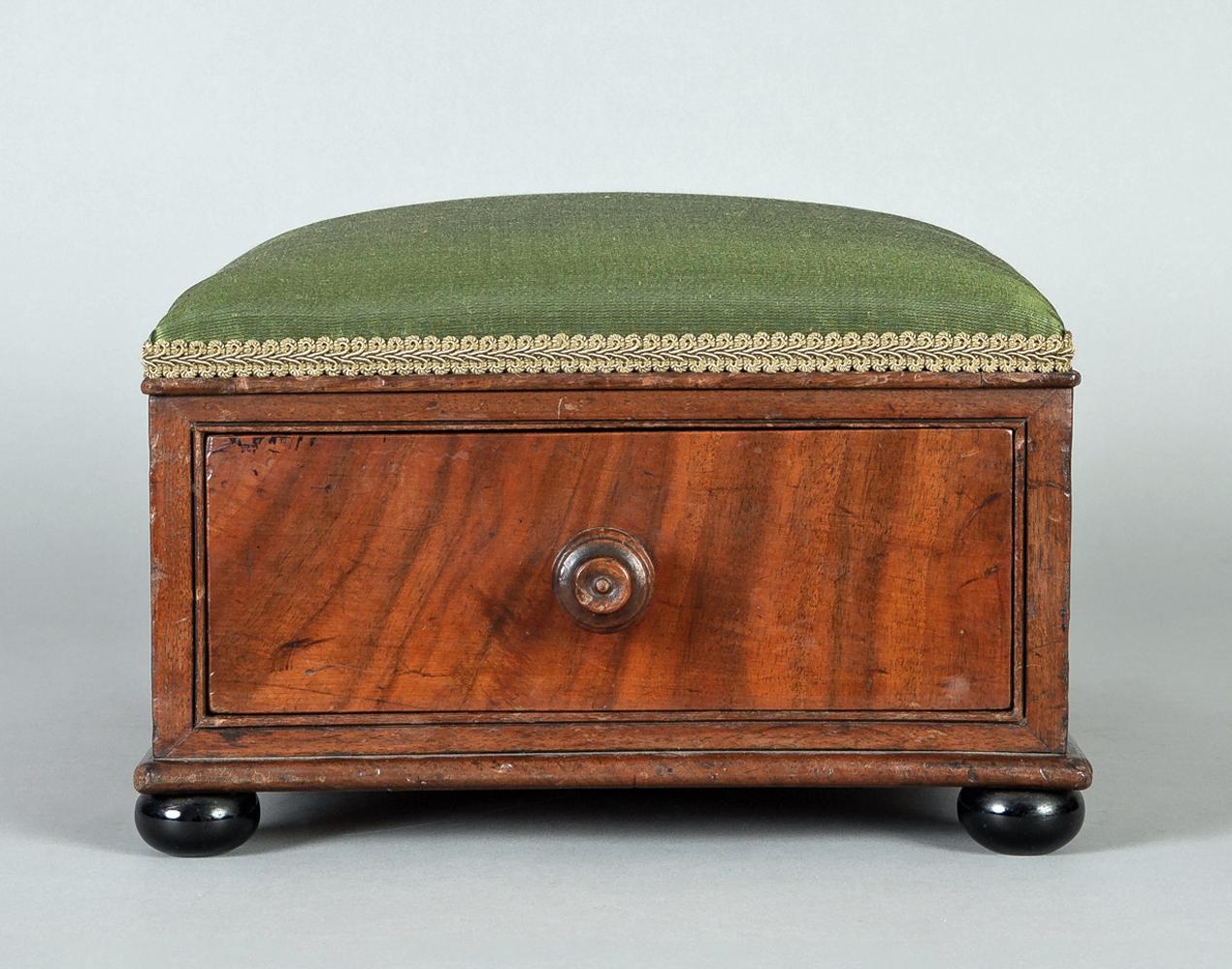 A Victorian mahogany footstool with a drawer that runs the depth of the stool with a turned wooded knob, covered in green silk and mounted on ebonized bun feet.