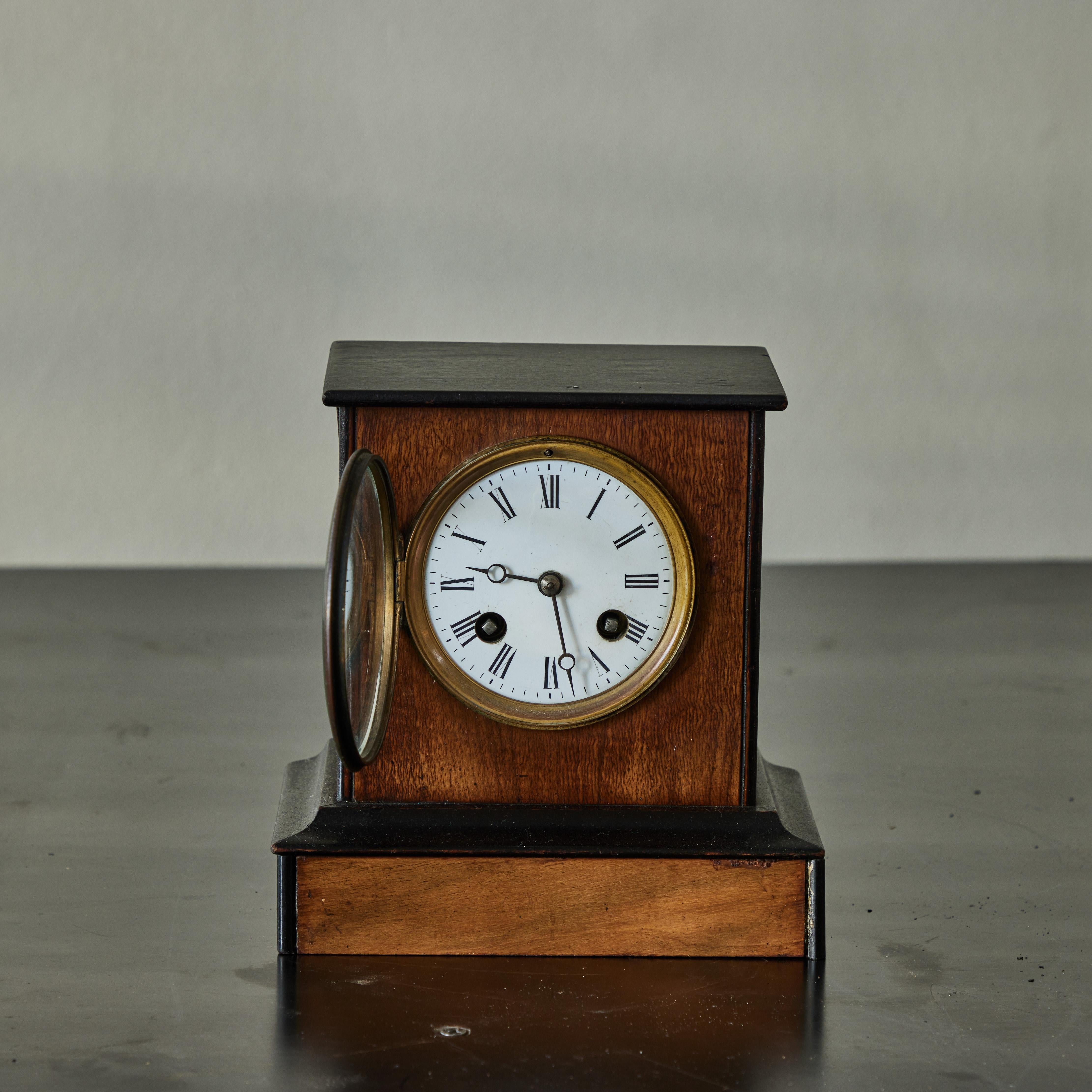 Decorative walnut wood mantle clock from Victorian England. Simple yet refined, the piece features a raised pedestal base, ebonized trim, and glass-fronted white enamel clock-face with delicate Roman numeral lettering. 

England, circa